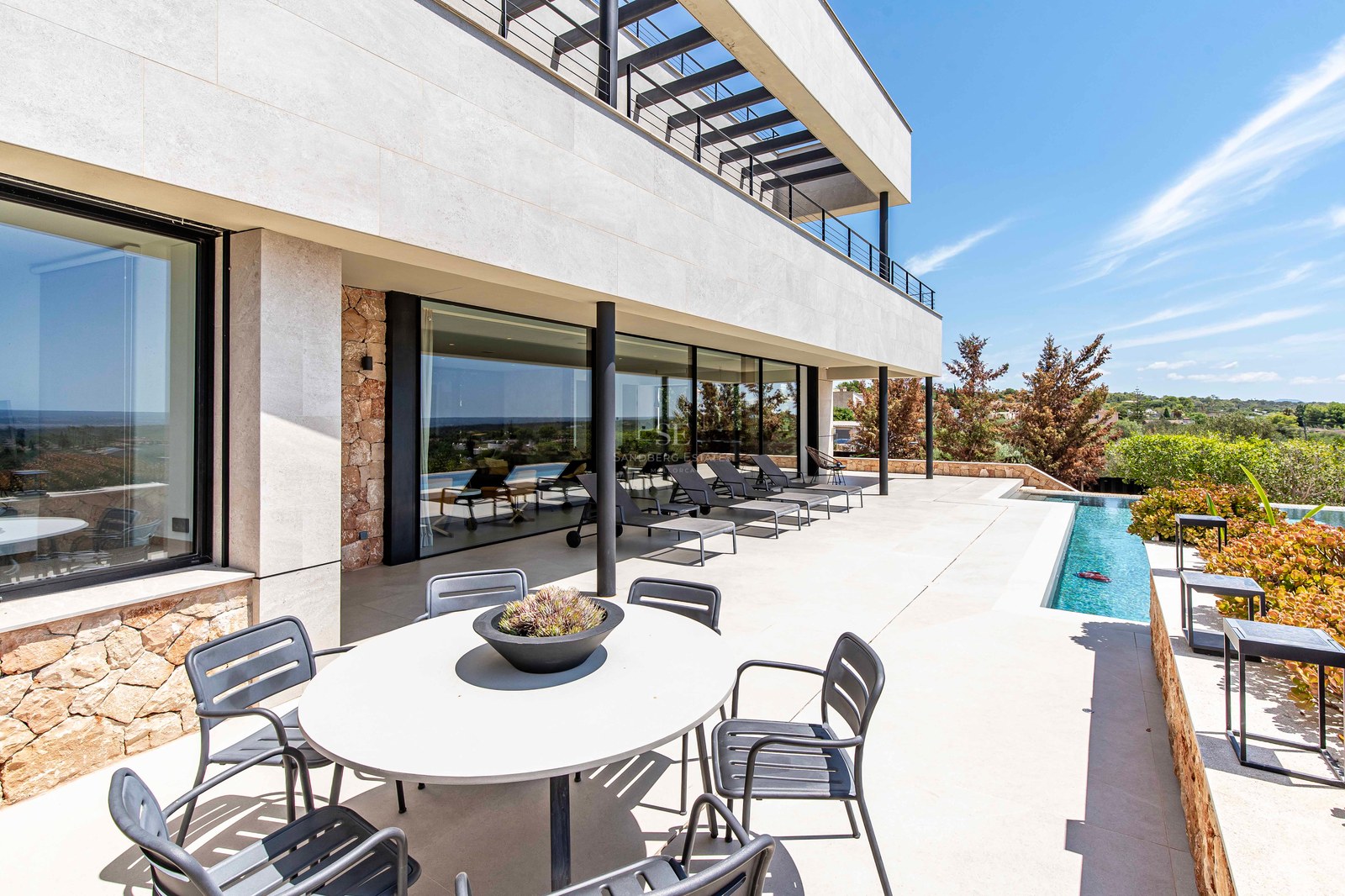 Sun-drenched terrace with grey loungers, outdoor dining area, and a sleek pool against a modern stone facade.
