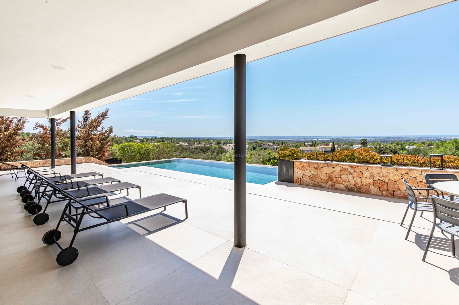A spacious modern white tiled terrace with black sun loungers, an infinity pool, and a view of the landscape and sea.