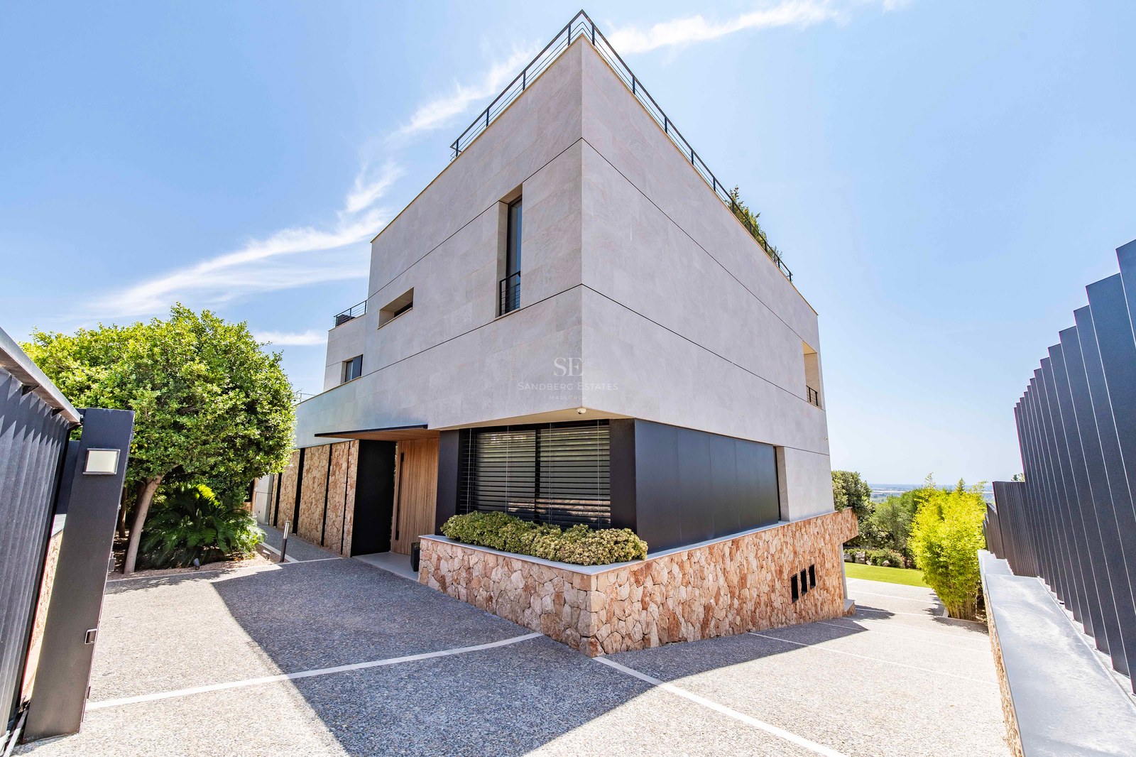 Modern villa exterior featuring natural stone walls, wooden entrance, and a gravel driveway under a clear blue sky.