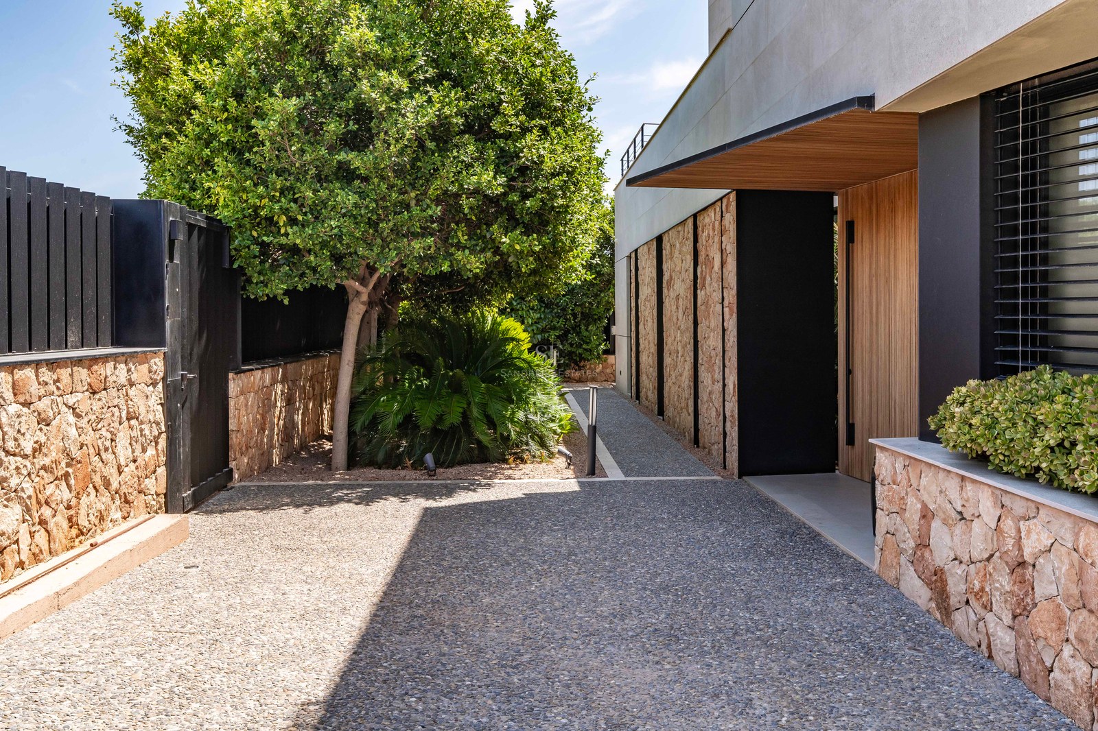 A contemporary villa entrance featuring a wooden door, stone walls, a gravel path, and lush green trees under bright sun.