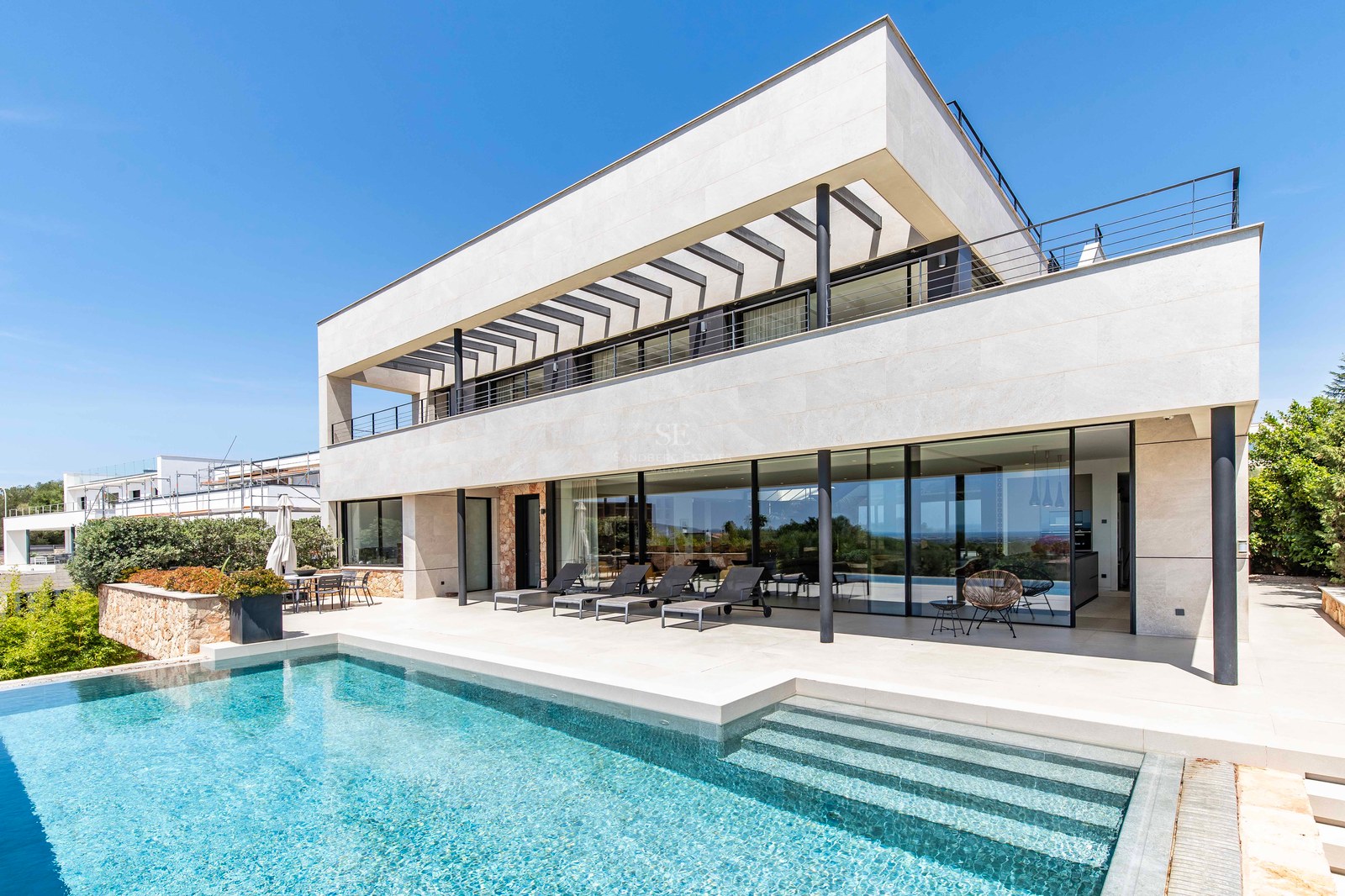 Luxury villa facade featuring a crystal-blue swimming pool, expansive glass windows, and stone cladding under a clear sky.