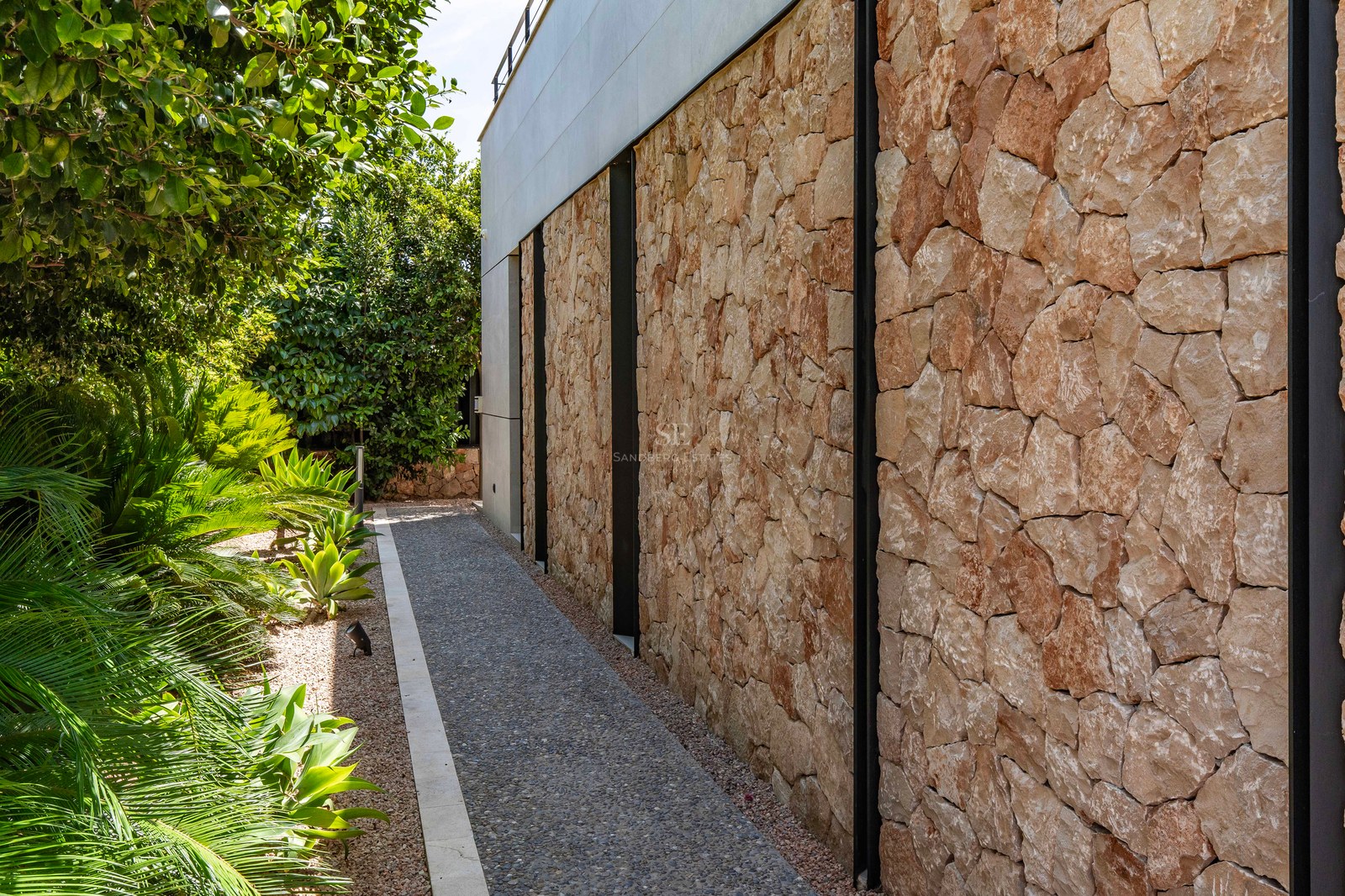 Side view of a contemporary villa's stone facade with a gravel path and lush green Mediterranean plants.