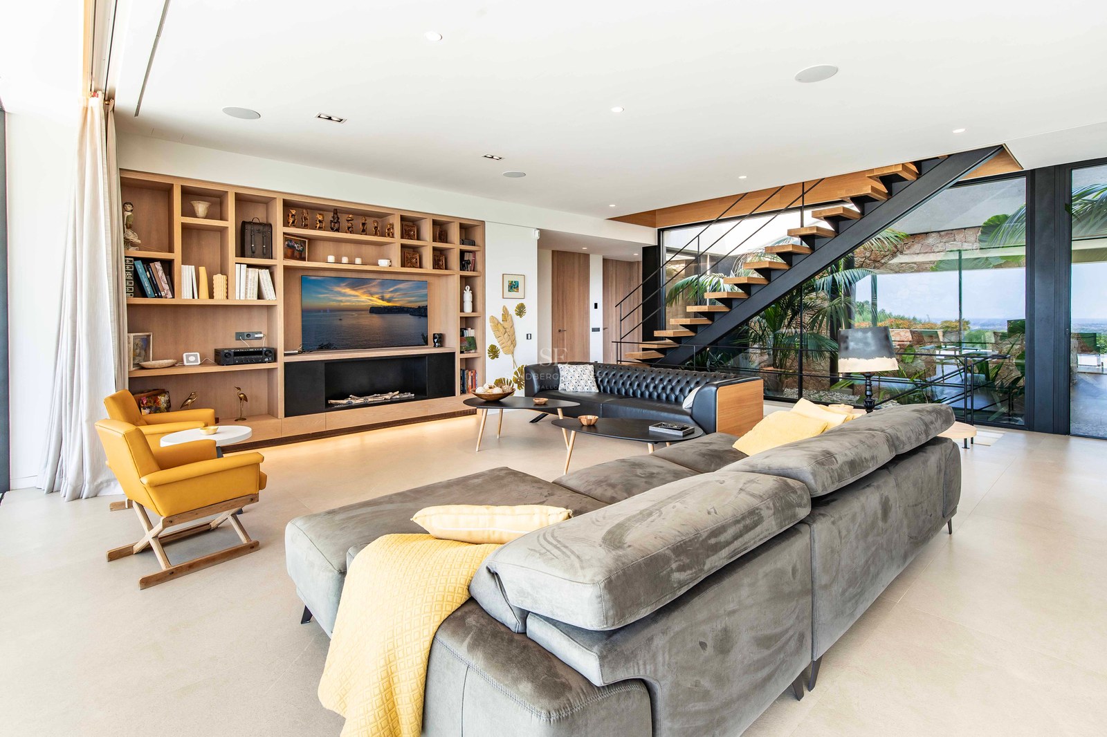 Modern living room featuring a grey sectional sofa, custom oak shelving, and a floating staircase with garden views.