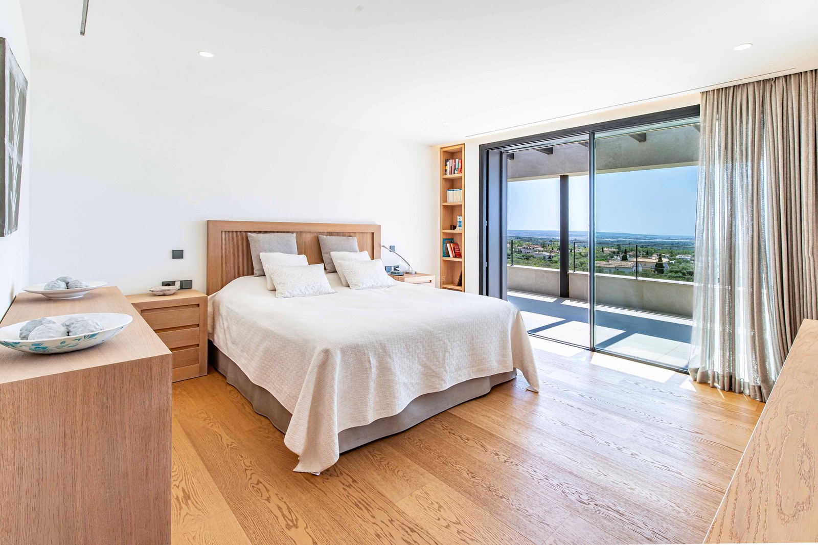 Bright modern bedroom with light oak floors and floor-to-ceiling glass doors opening to a sunny terrace and sea views.