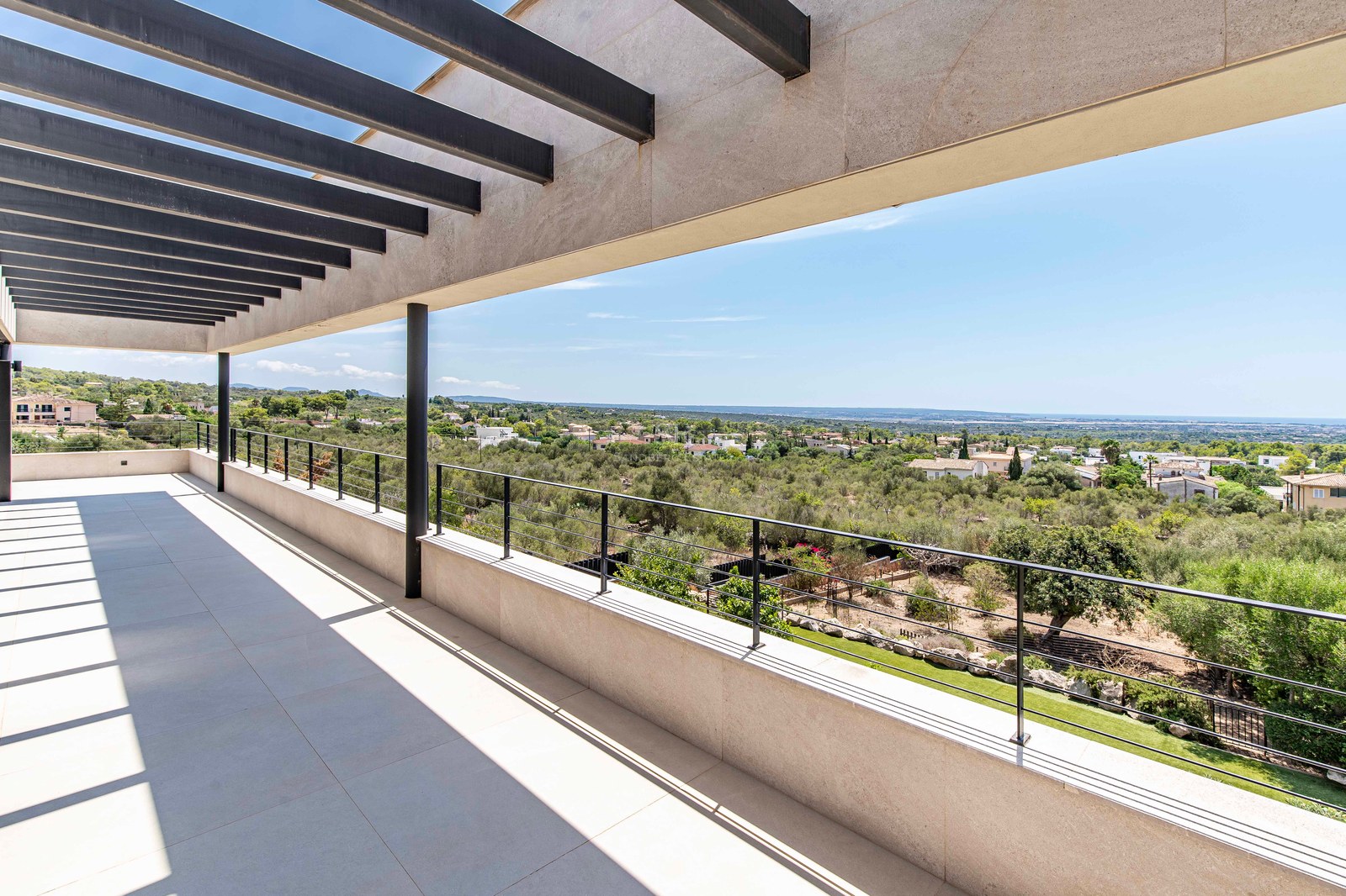 Wide modern terrace with a black slat pergola overlooking a green valley and distant coastline under a clear blue sky.