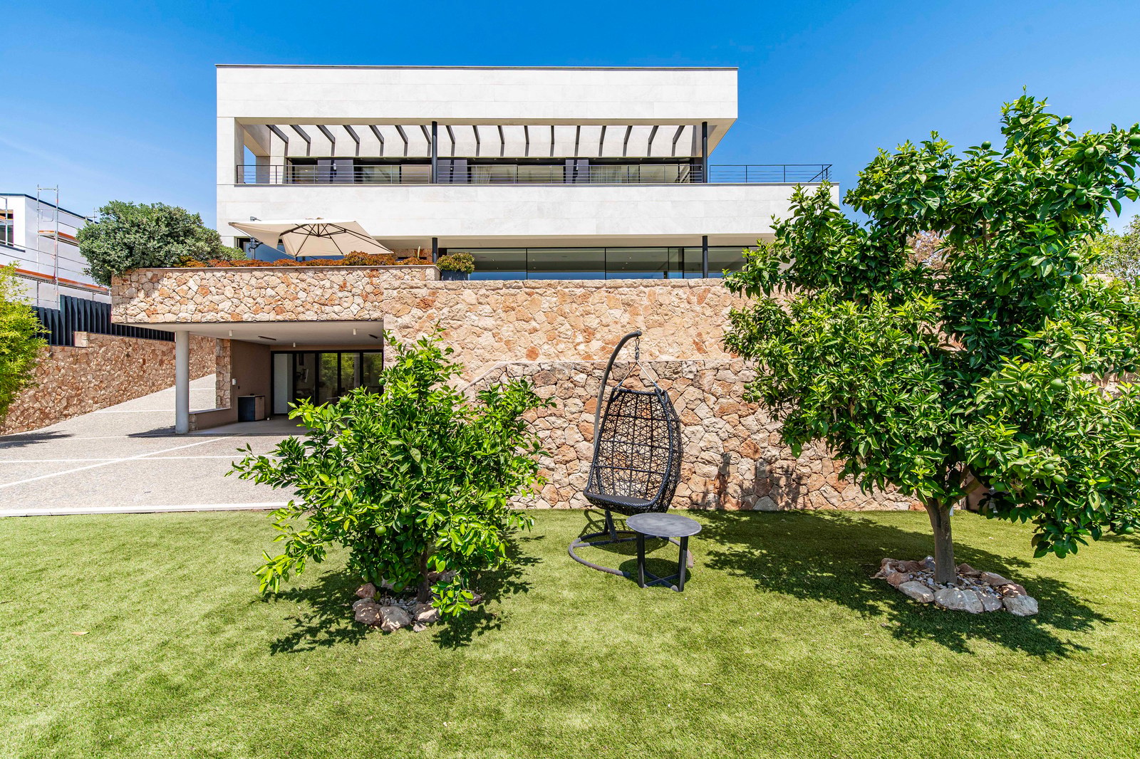 Exterior view of a modern three-level villa featuring stone walls, white upper levels, glass balconies, and a green lawn.