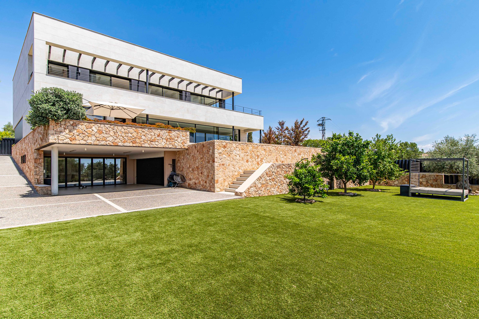 Modern white villa with natural stone walls, a large green lawn, and minimalist landscaping under a clear blue sky.