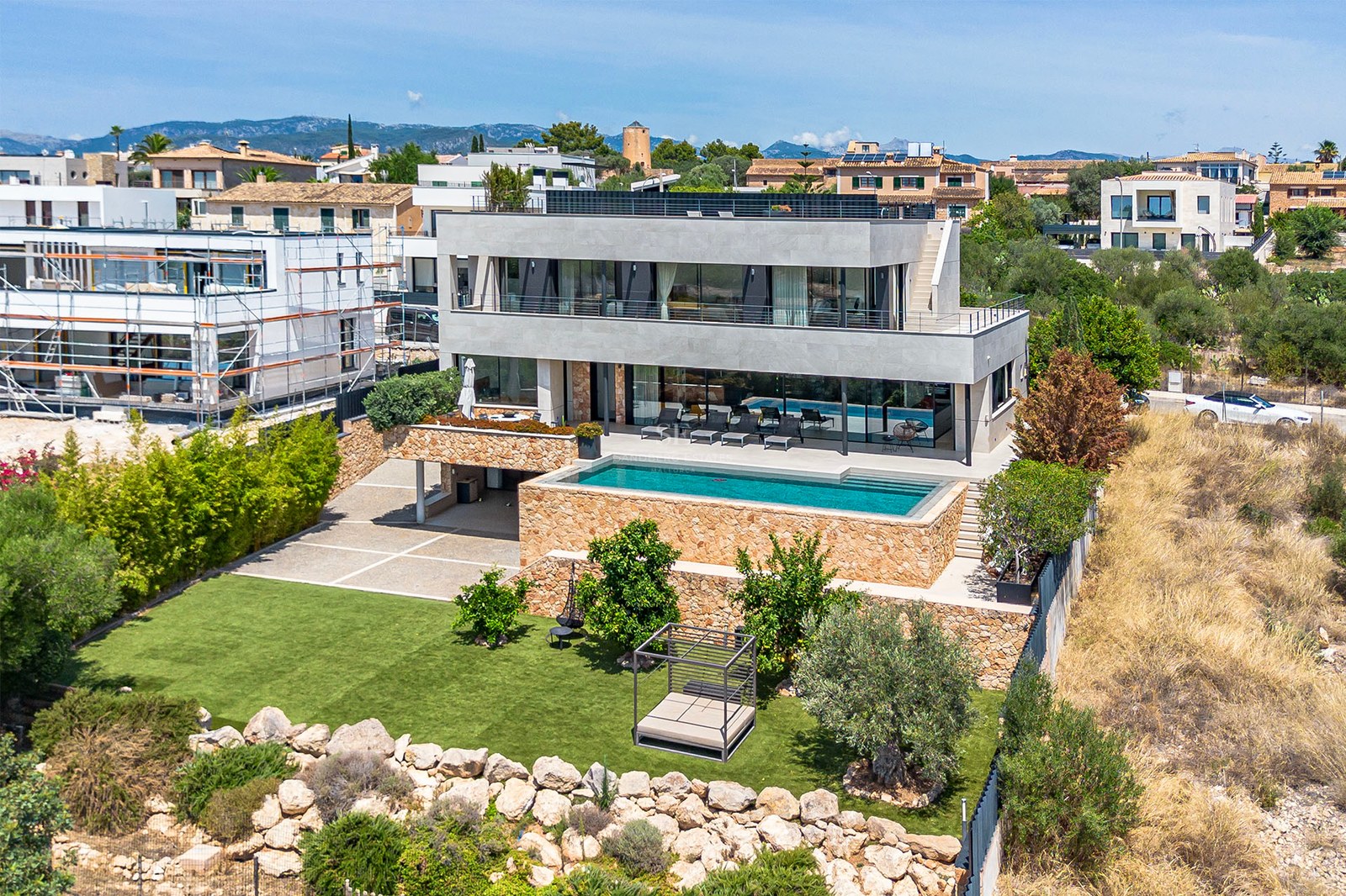 Aerial view of a contemporary three-story villa with a turquoise pool, natural stone walls, and a manicured lawn garden.