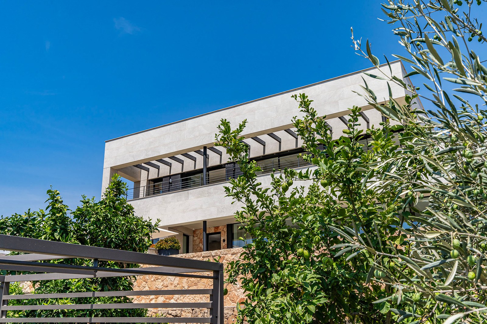 Modern two-story villa with stone facade, glass balconies, and lush olive and citrus trees under a clear blue sky.