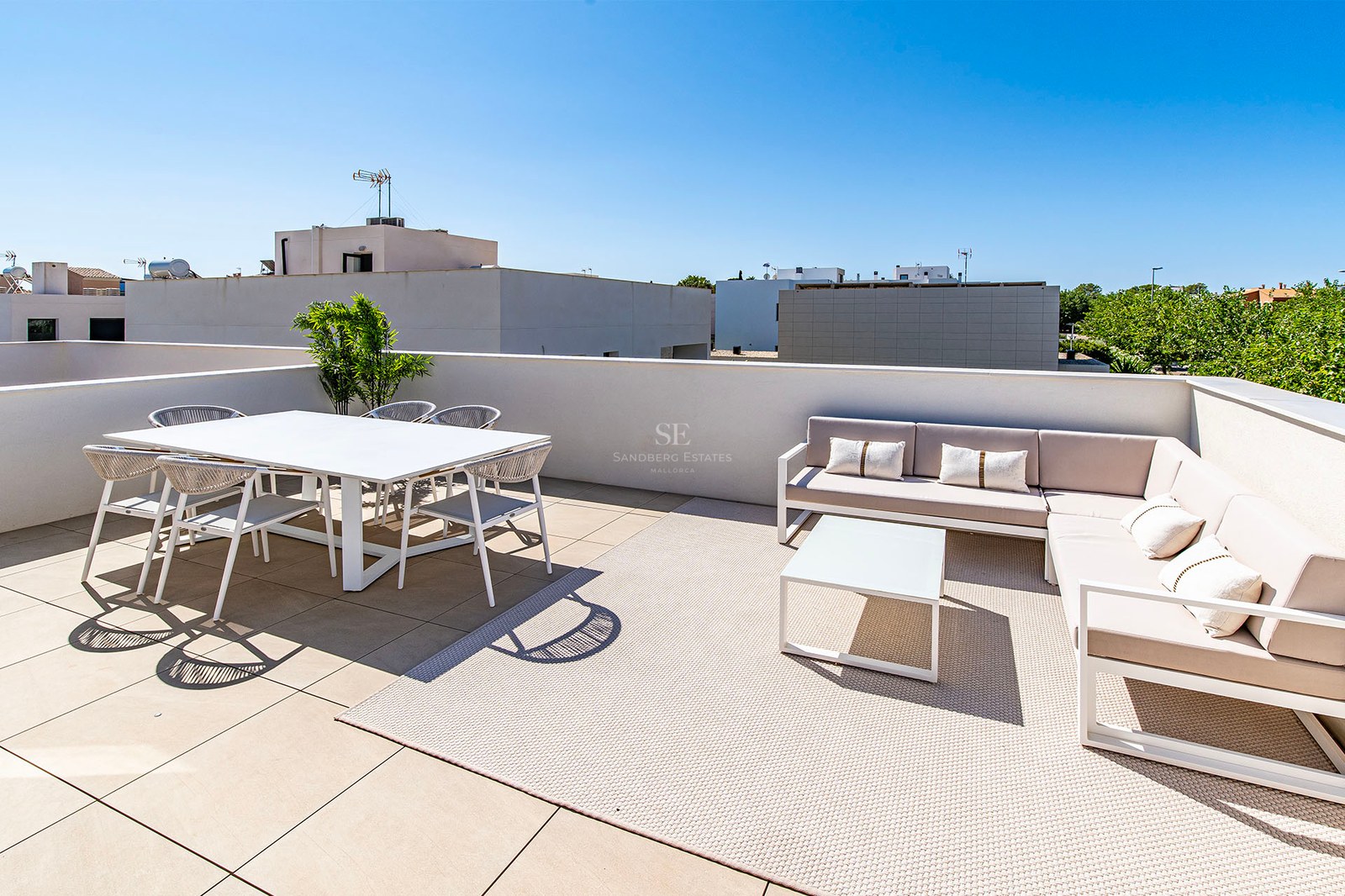 Terraza soleada con mesa de comedor blanca, sillas de cuerda y sofá rinconero beige sobre alfombra de exterior.