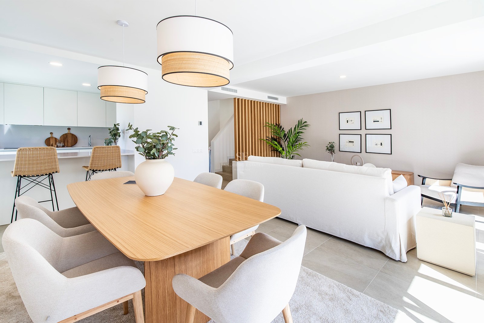 Salón comedor luminoso con mesa de madera clara, sillas beige y sofá blanco bajo luz natural.