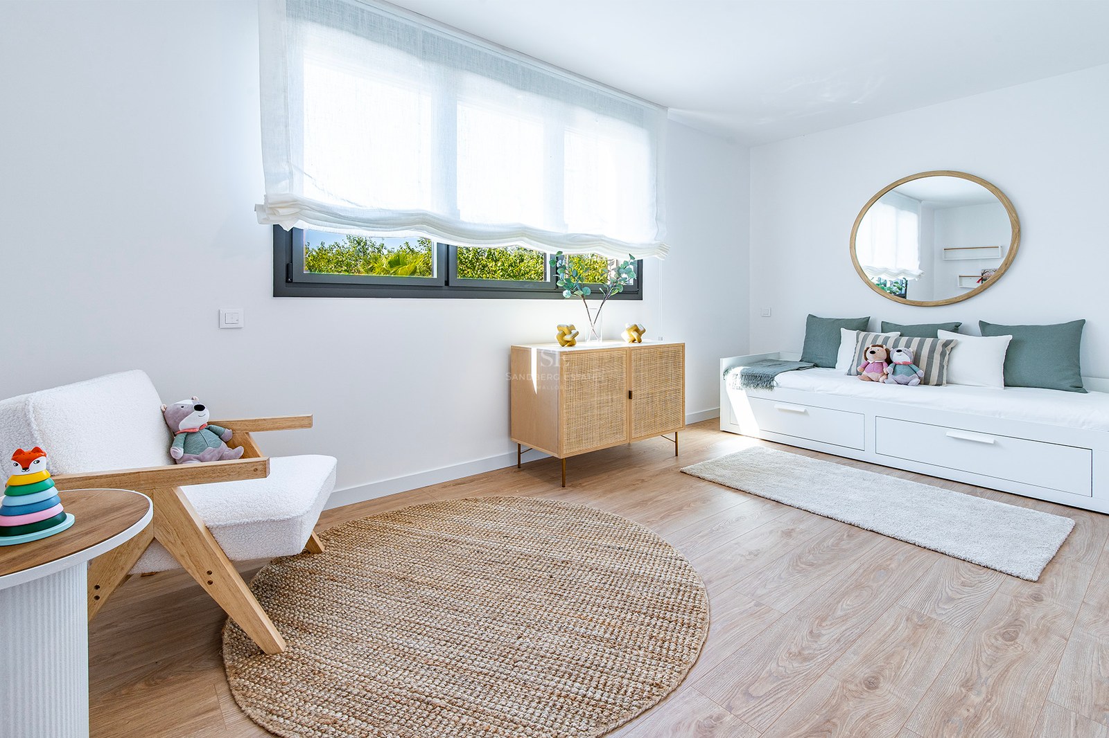 Habitación moderna con suelos de roble, cama nido blanca, alfombra de yute y aparador de ratán bajo una ventana luminosa.