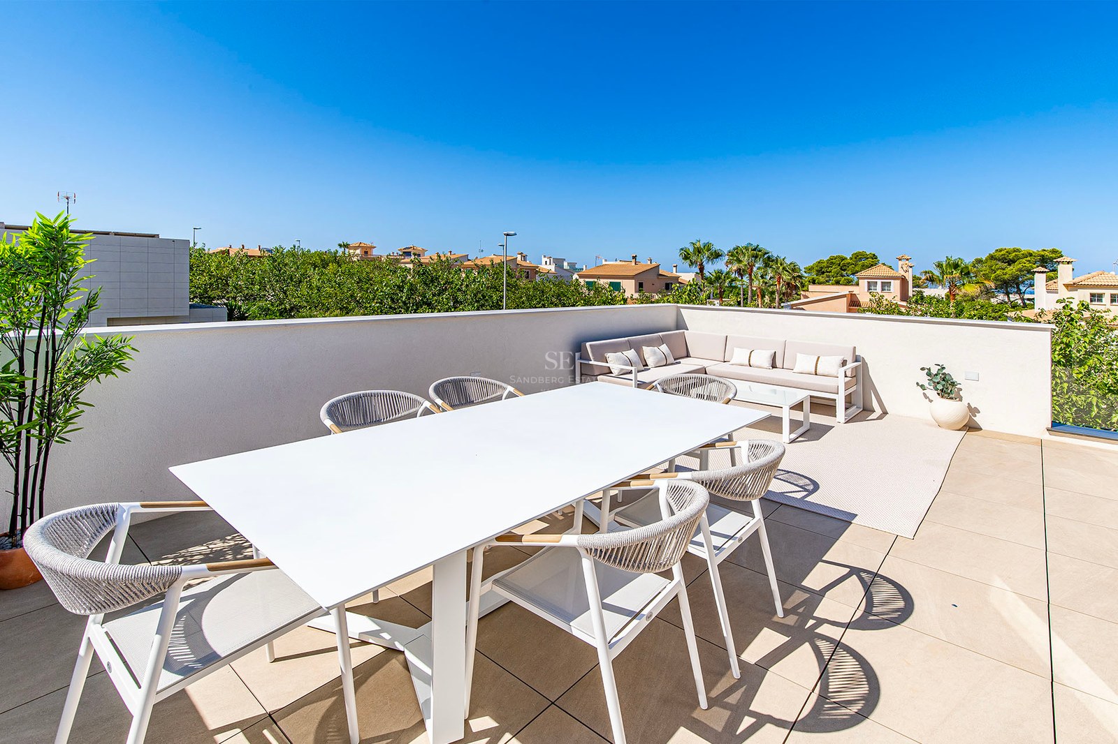 Terraza luminosa en la azotea con mesa de comedor blanca y zona de estar bajo un cielo azul despejado.