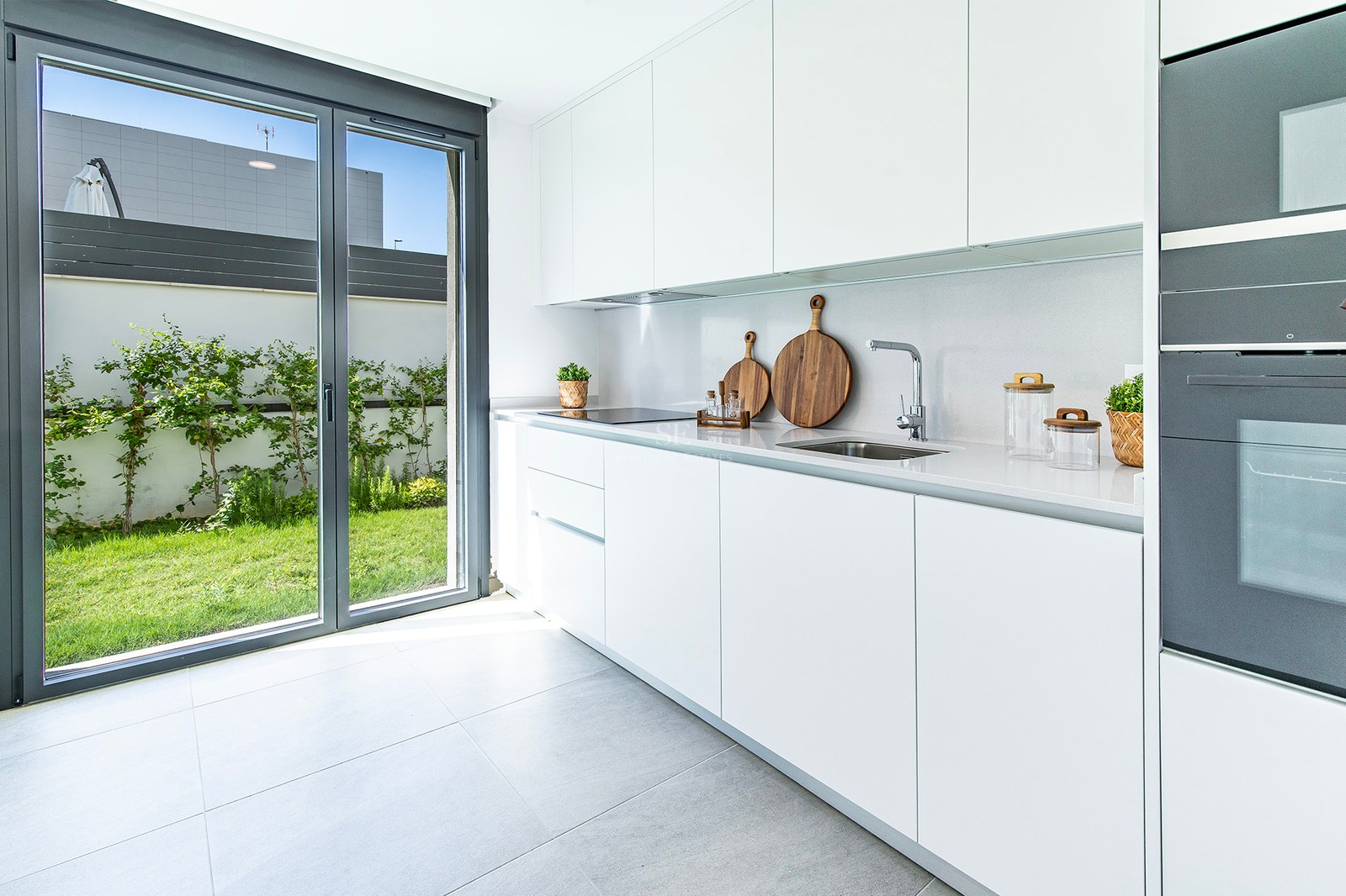Cocina blanca con armarios sin tiradores, encimera de piedra y puerta acristalada hacia un jardín privado.