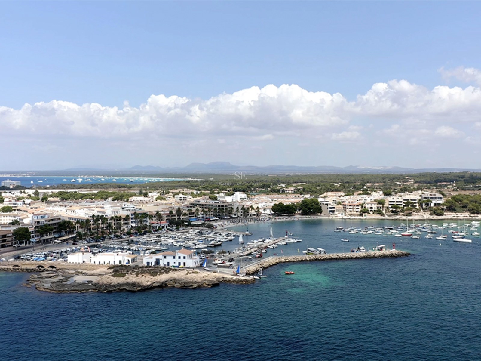 Elevated aerial view of a busy marina with white yachts, coastal buildings, and clear turquoise sea under a blue sky.