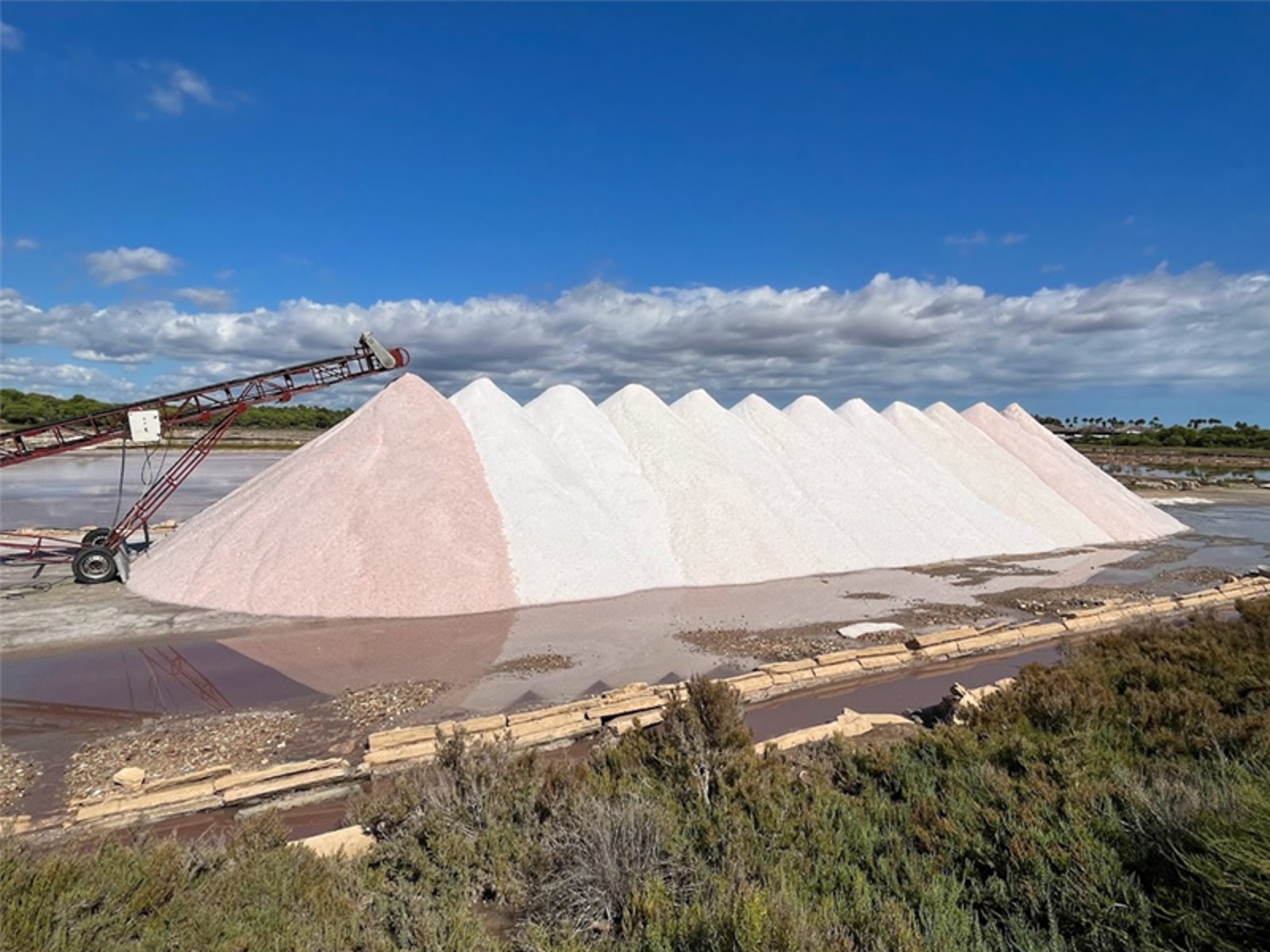 Large white and pink salt mounds under a clear blue sky with a conveyor belt at a coastal salt flat in Mallorca.