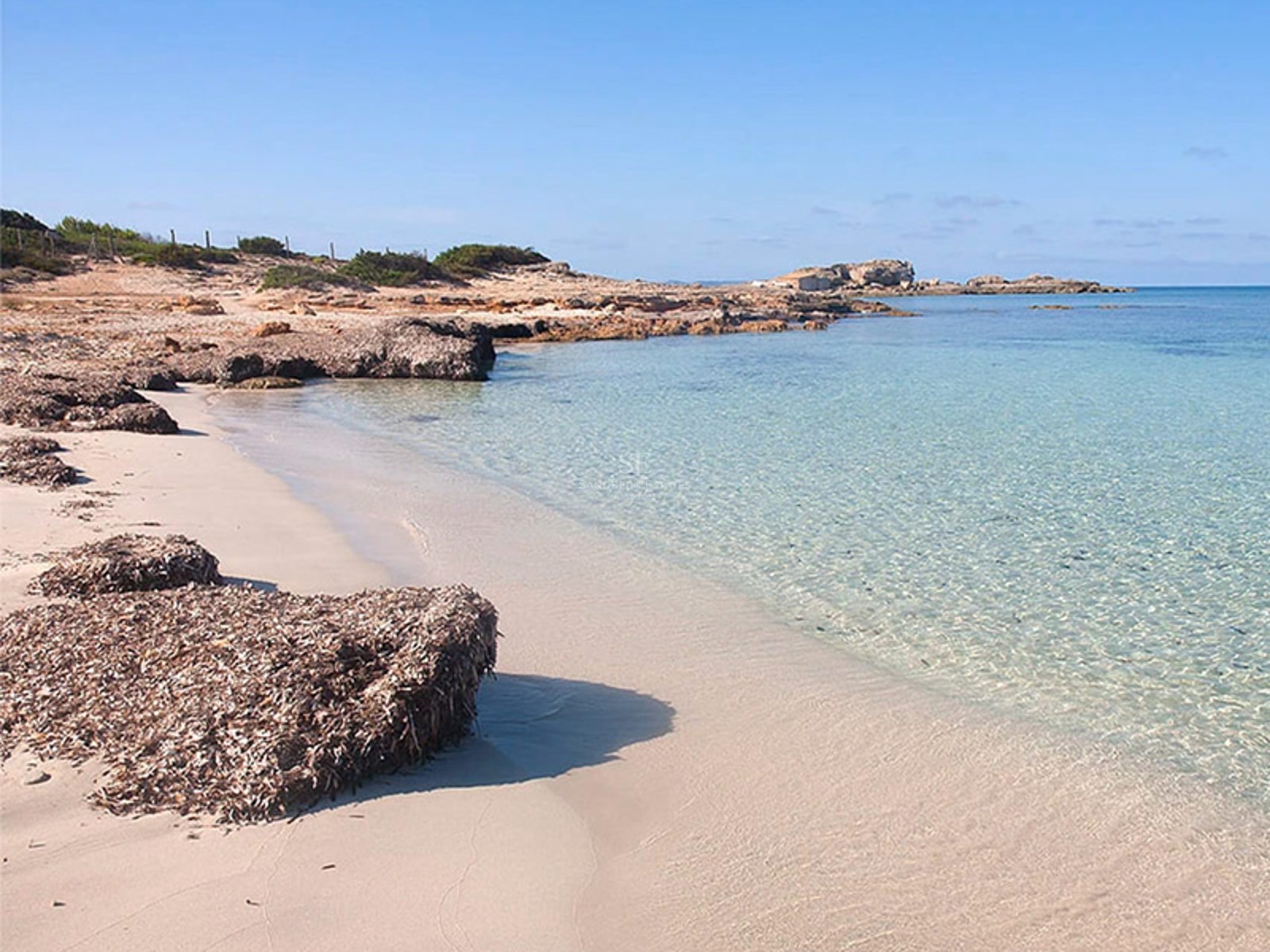 A quiet sandy beach with clear turquoise water and rocky outcrops under a clear blue sky.