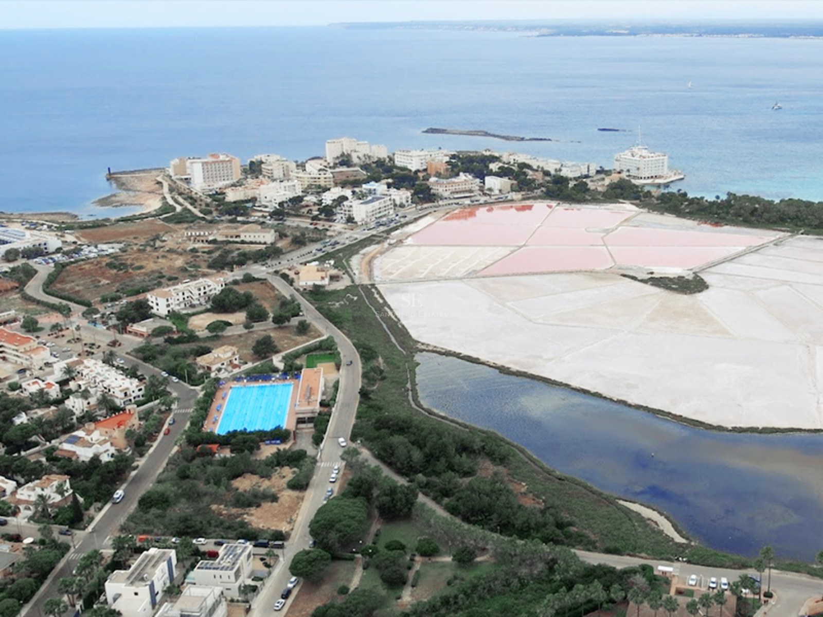 High-angle aerial view of a coastal Mediterranean town with pink salt flats, turquoise sea, and urban buildings.