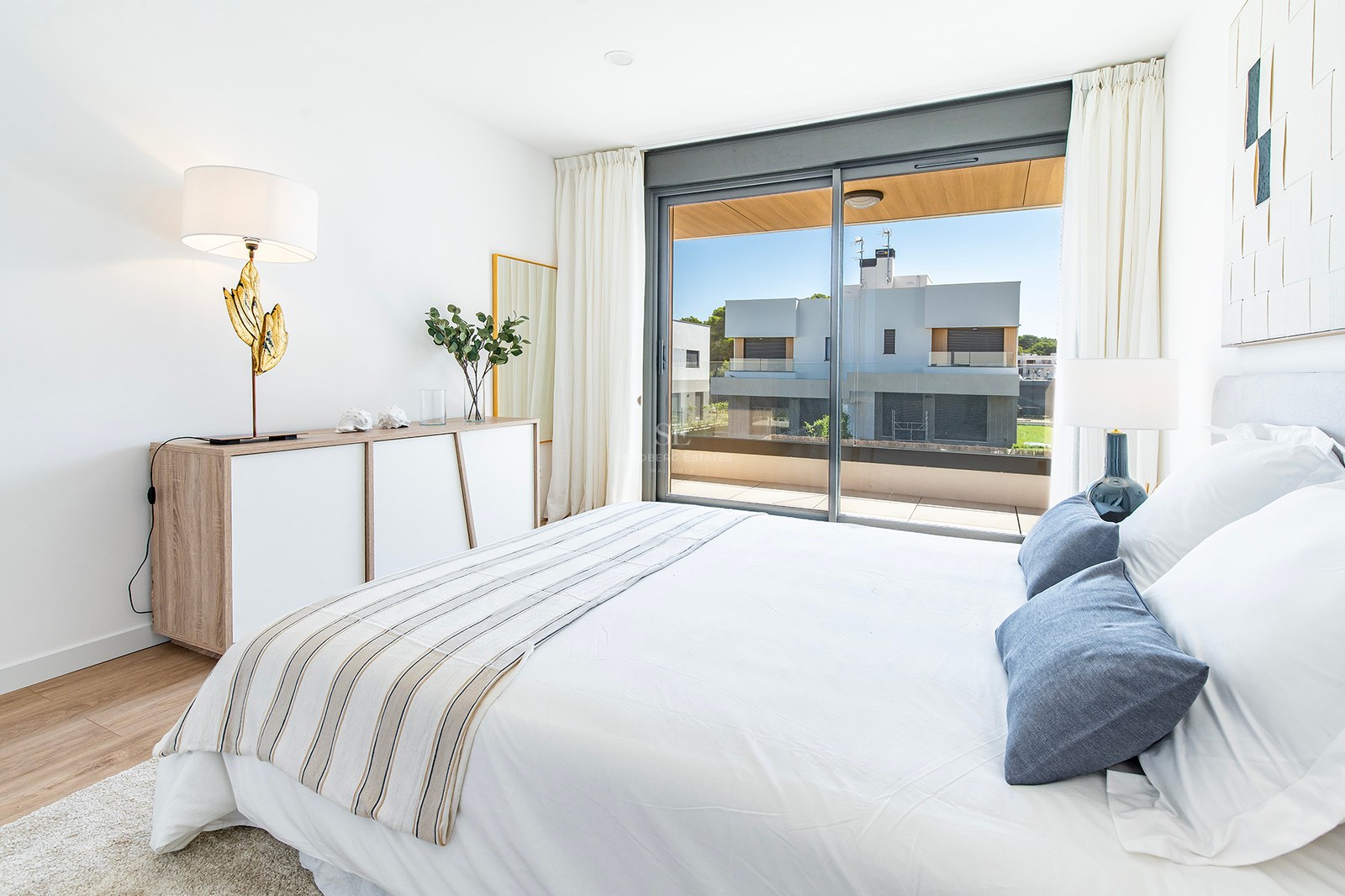 Helles Schlafzimmer mit Doppelbett, Parkettboden und großen Glasschiebetüren zu einer sonnigen Terrasse.