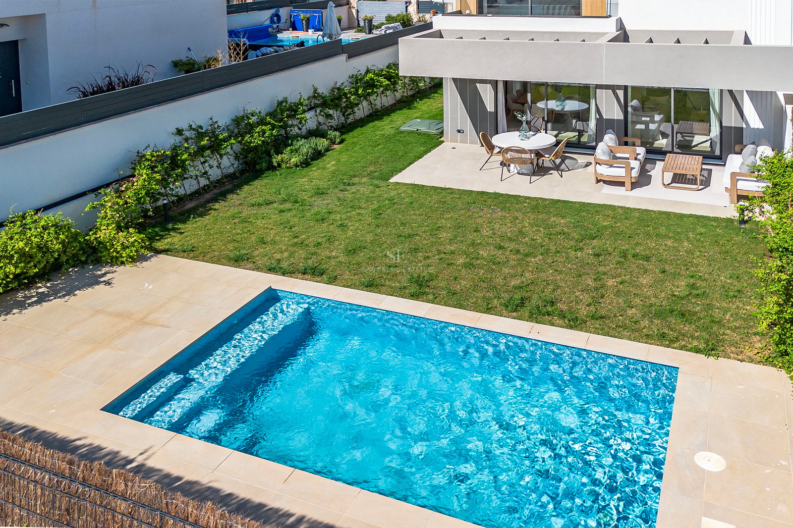 Draufsicht auf einen rechteckigen blauen Pool, Rasenfläche und eine moderne Terrasse mit weißen Gartenmöbeln.