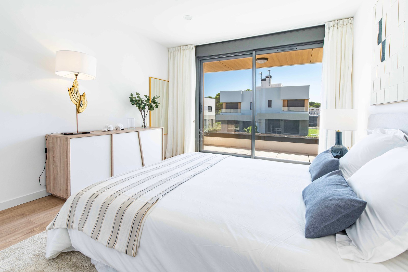 Modernes Schlafzimmer mit weißem Bett, blauen Kissen, Holzboden und großen Glastüren zur Terrasse.