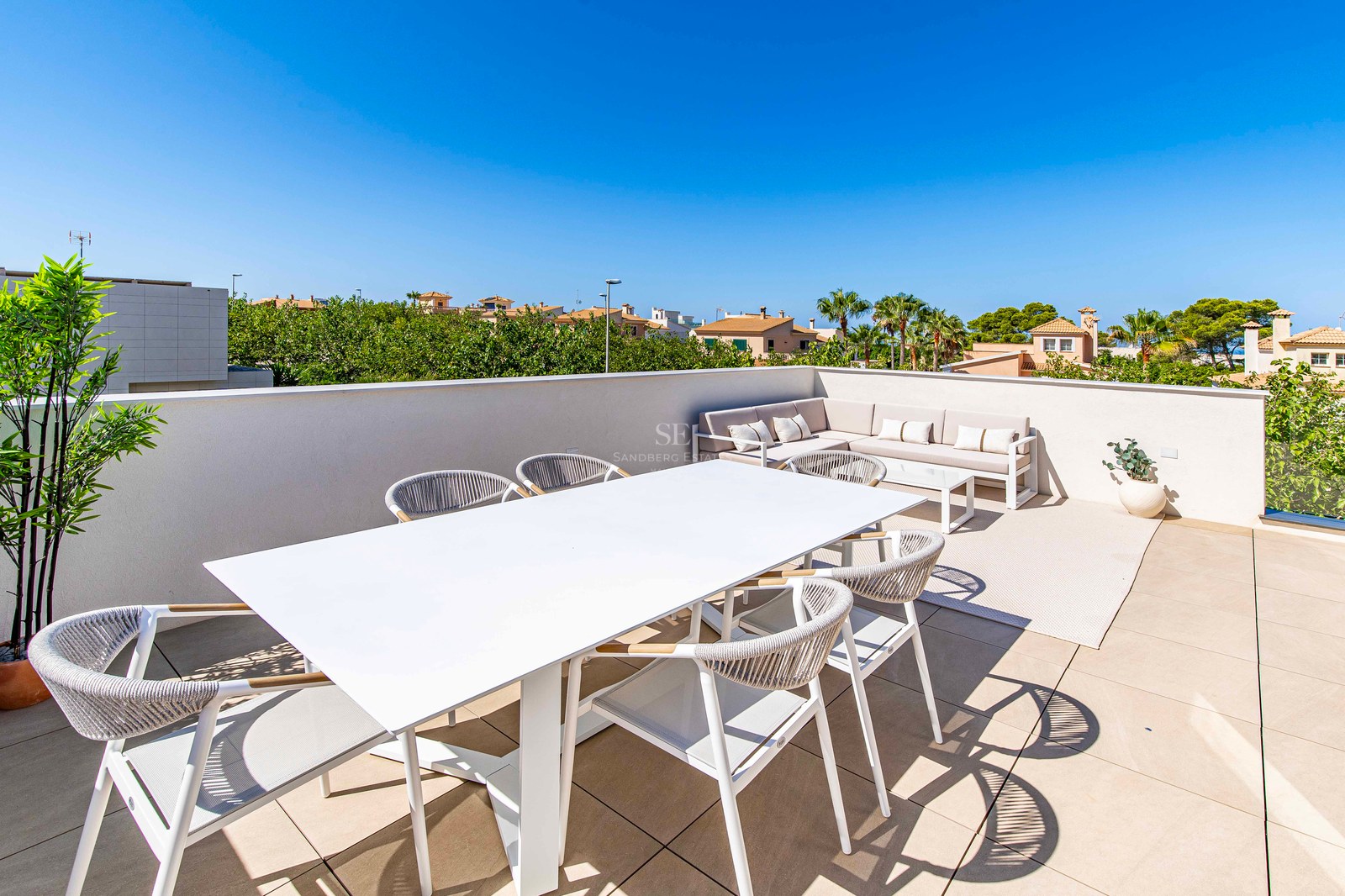 Sonnige Dachterrasse mit weißem Esstisch, Seilstühlen und Ecksofa unter blauem Himmel.