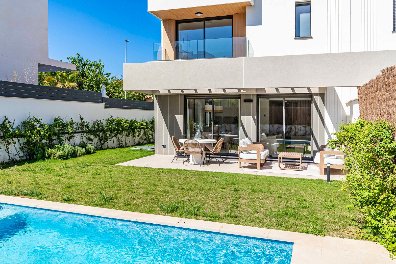 Moderne weiße Villa mit türkisfarbenem Pool, grünem Rasen und möblierter Terrasse unter blauem Himmel.