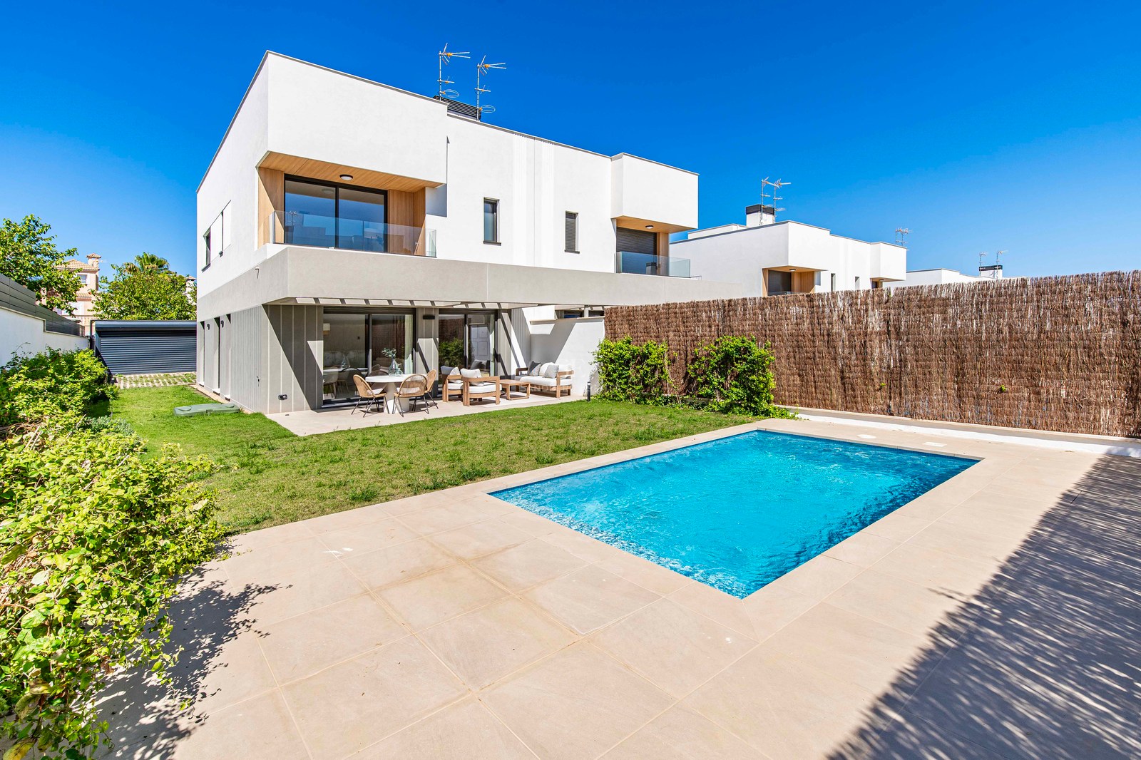 Außenansicht einer modernen Villa mit blauem Pool, Rasenfläche und möblierter Terrasse unter blauem Himmel.