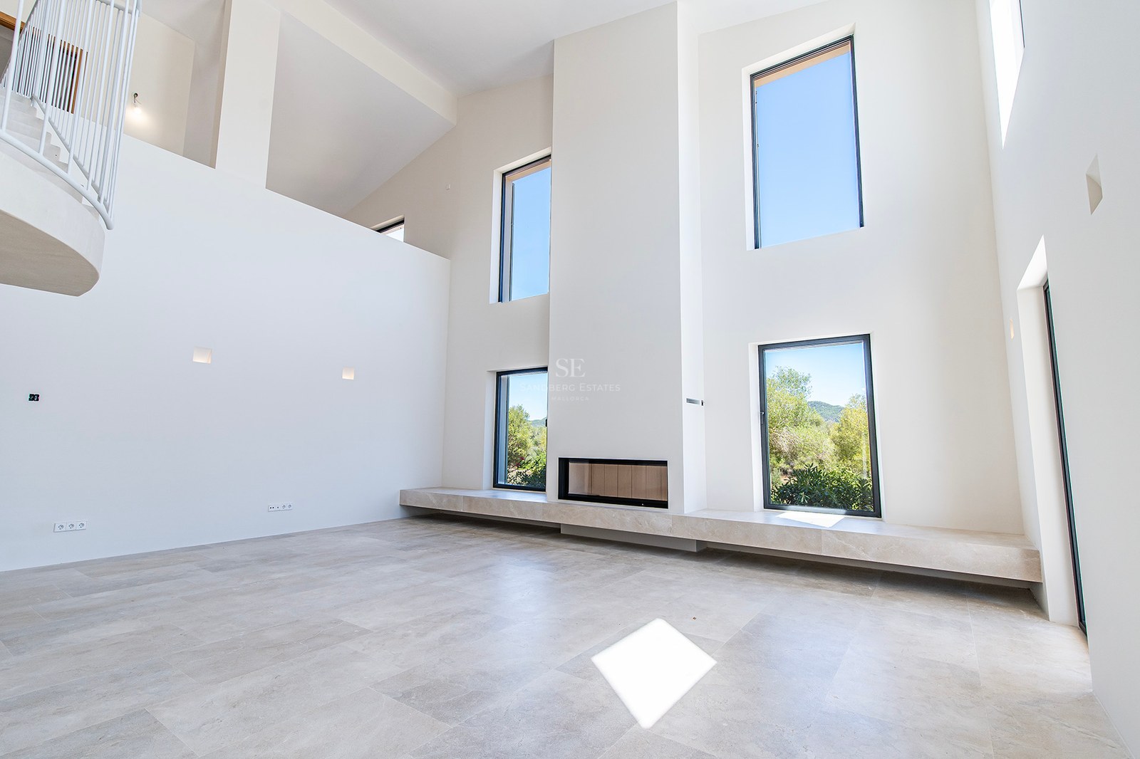 Salón minimalista de doble altura con paredes blancas, grandes ventanales, chimenea integrada y suelos de piedra clara.