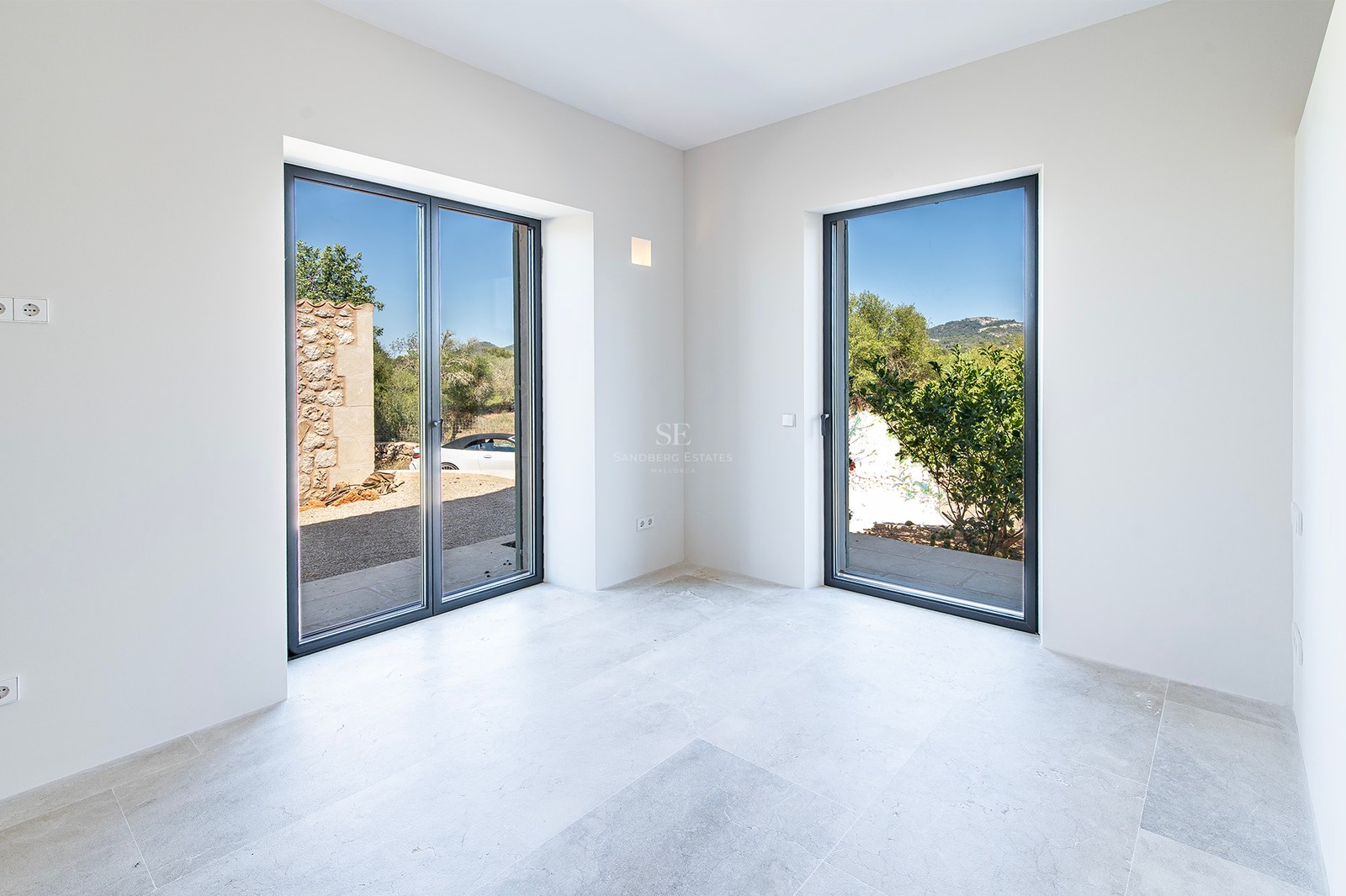 Habitación minimalista vacía con paredes blancas, suelos de piedra gris y dos grandes ventanales con vistas al exterior.