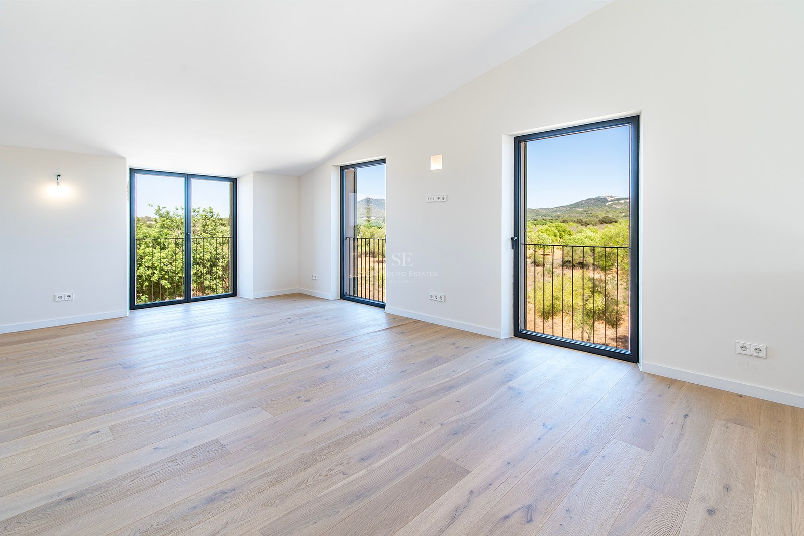 Habitación vacía con suelos de madera clara, techos inclinados y grandes ventanales con vistas al paisaje verde.