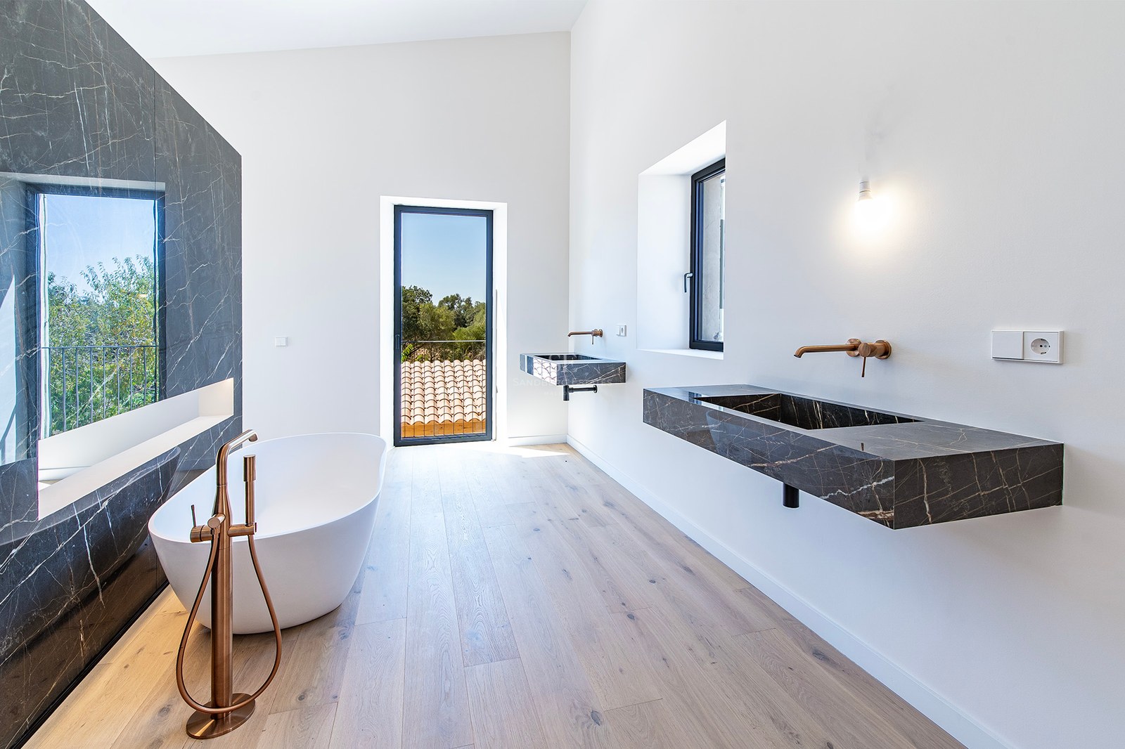 Baño moderno con bañera exenta, pared de mármol oscuro, lavabos suspendidos y grifería de bronce hacia el exterior.