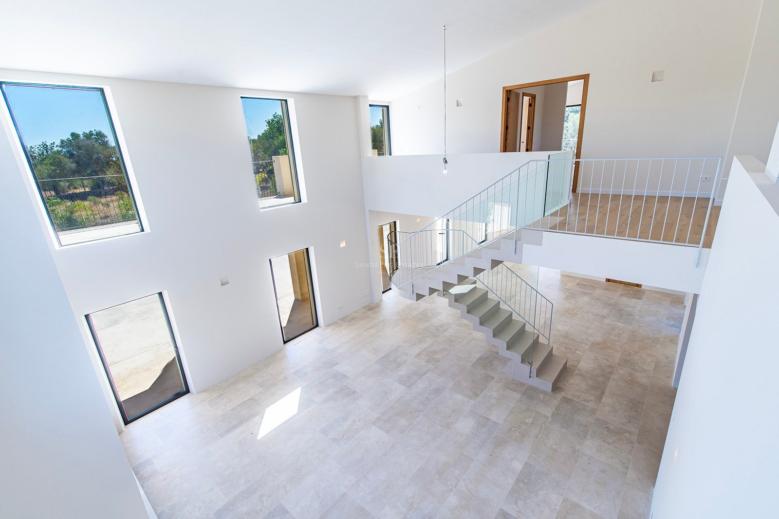 Salón de doble altura con suelos de piedra, escalera moderna y grandes ventanales con luz natural en villa de lujo.