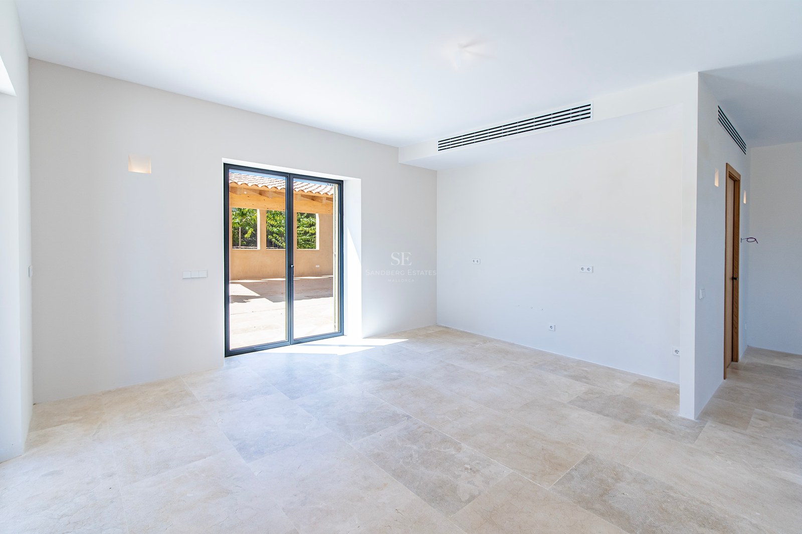 Habitación vacía con suelo de piedra natural crema, paredes blancas y puerta de cristal hacia un patio soleado.