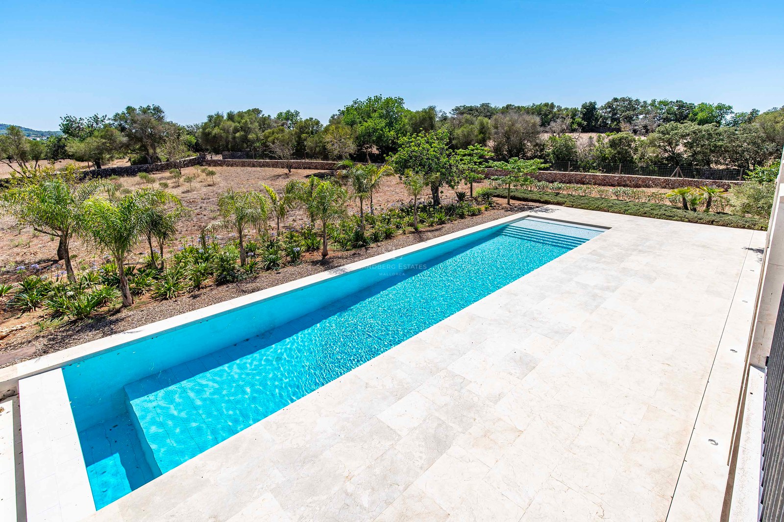 Piscina rectangular turquesa rodeada por una amplia terraza de piedra crema y vegetación mediterránea.