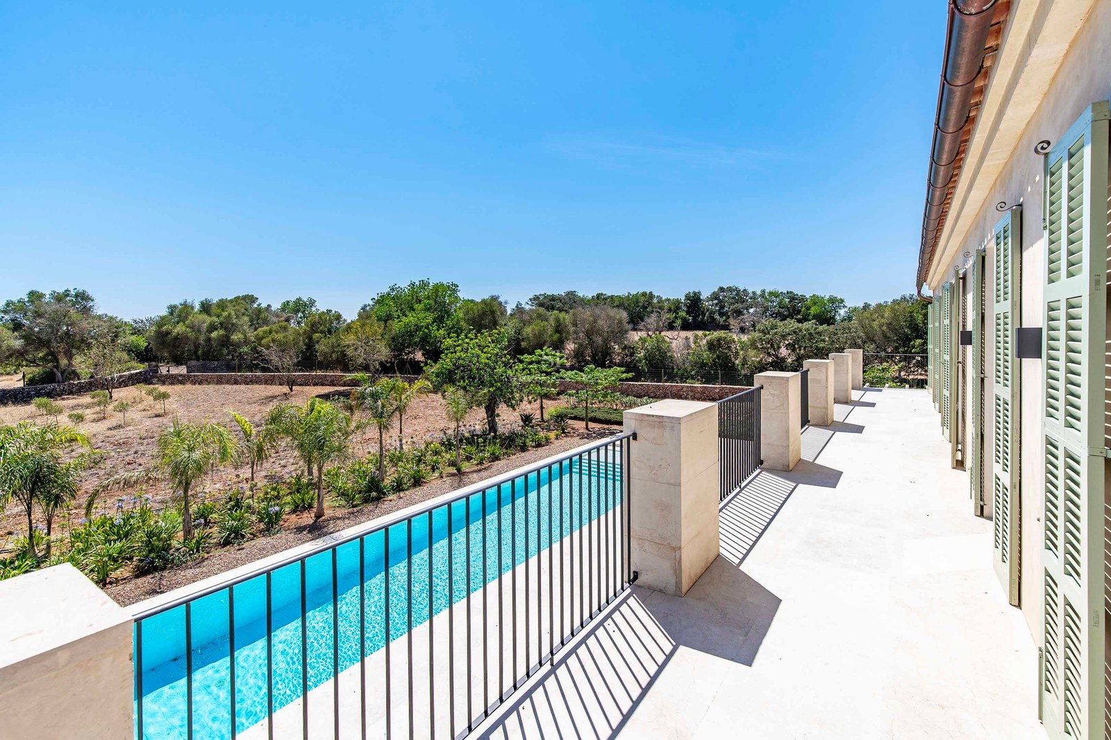 Vista desde terraza de piedra con barandilla negra hacia una piscina alargada y paisaje mediterráneo bajo cielo despejado.