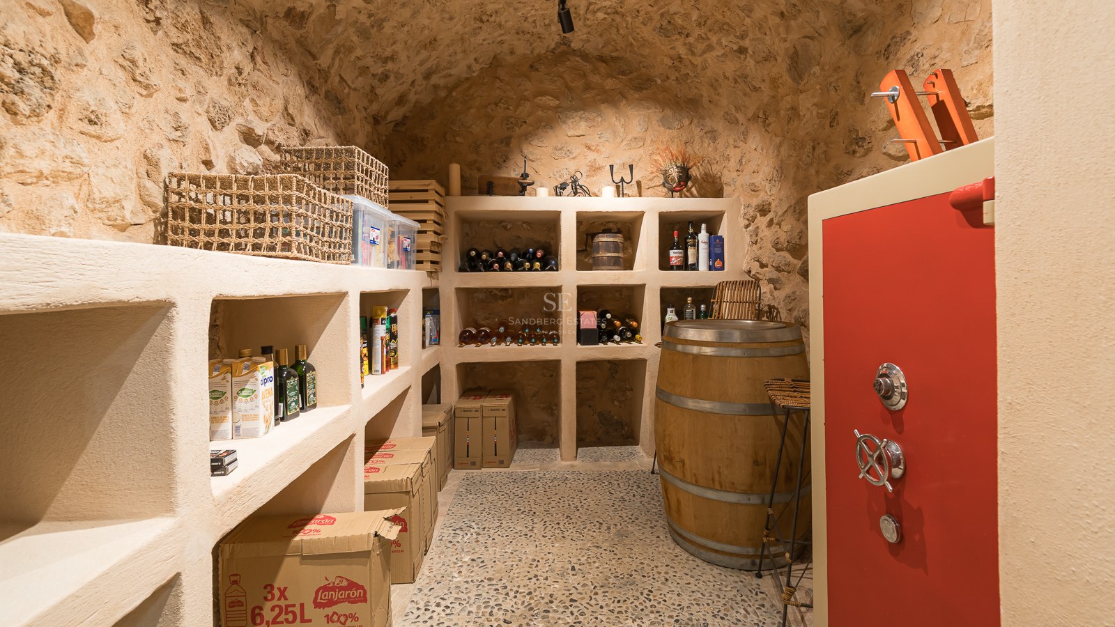 Bodega rústica con techos abovedados de piedra, estanterías integradas, una mesa de barril de madera y una caja fuerte roja.