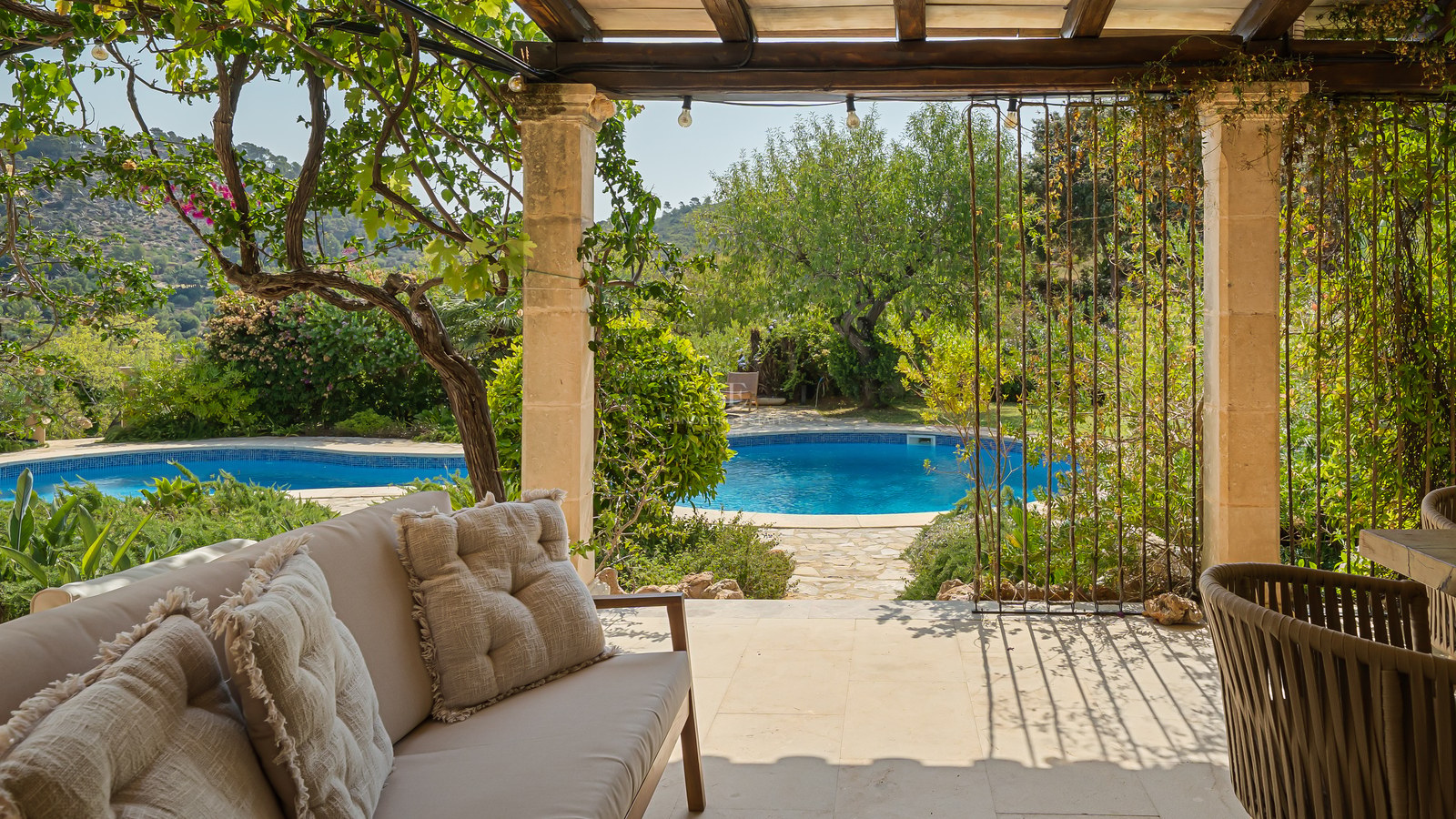 Covered stone terrace with comfortable sofa overlooking a circular blue pool surrounded by lush green gardens.