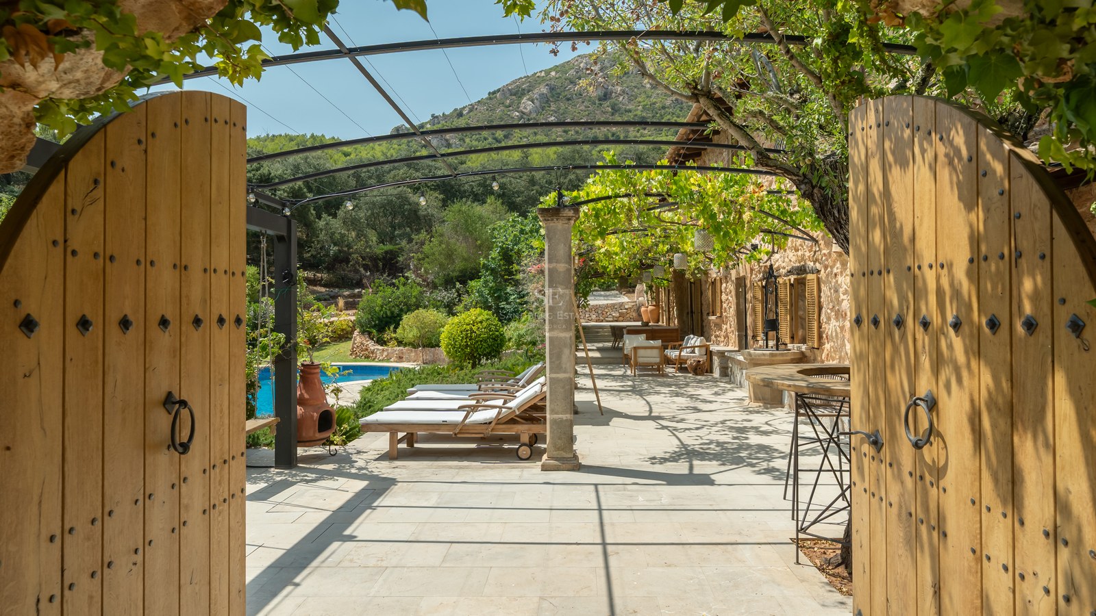 View through open wooden gates of a stone-paved terrace with sun loungers, vine-covered pergola, and pool.