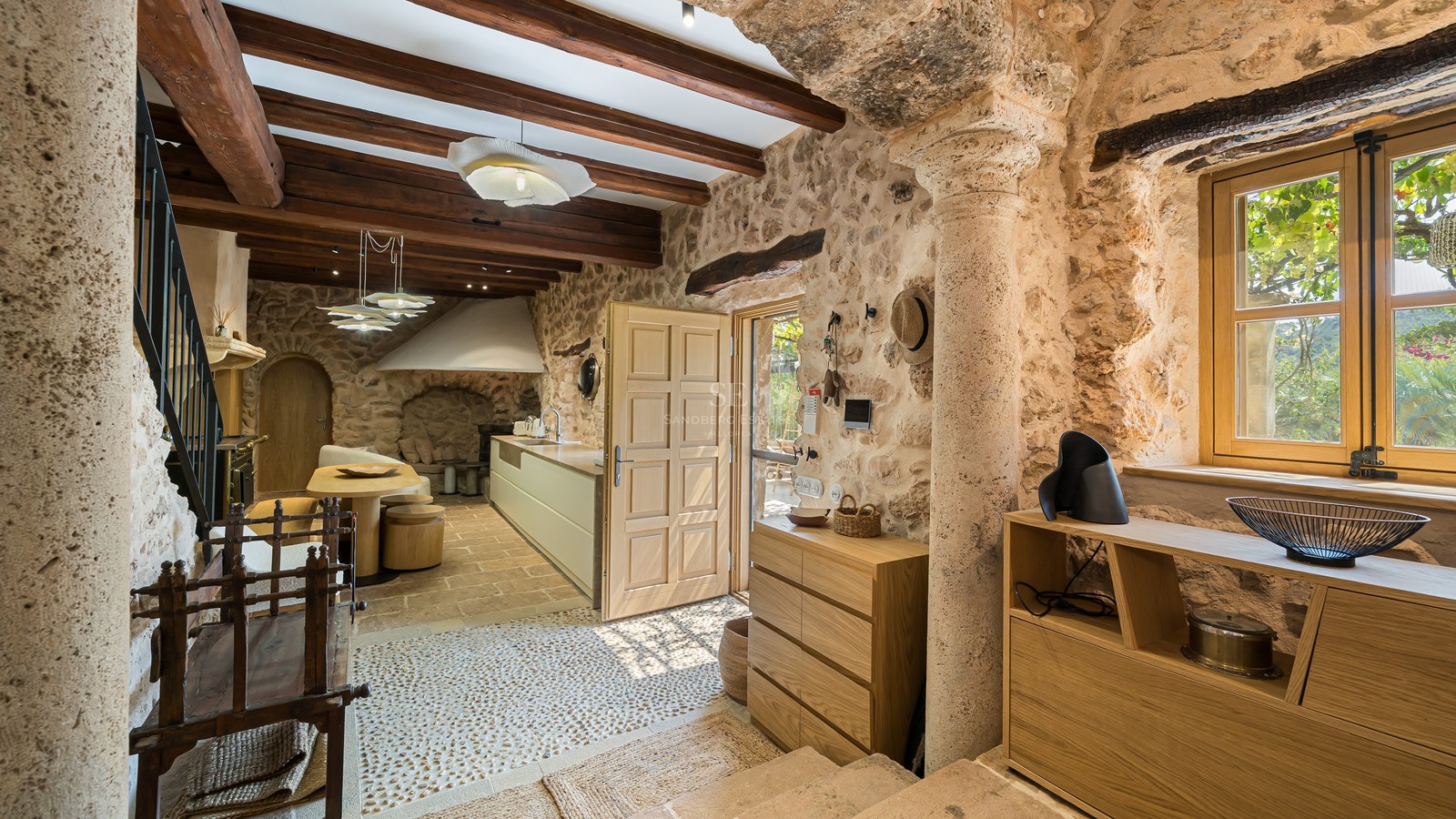 Interior view of a rustic foyer with stone walls, wooden ceiling beams, a stone column, and a view into a modern kitchen.