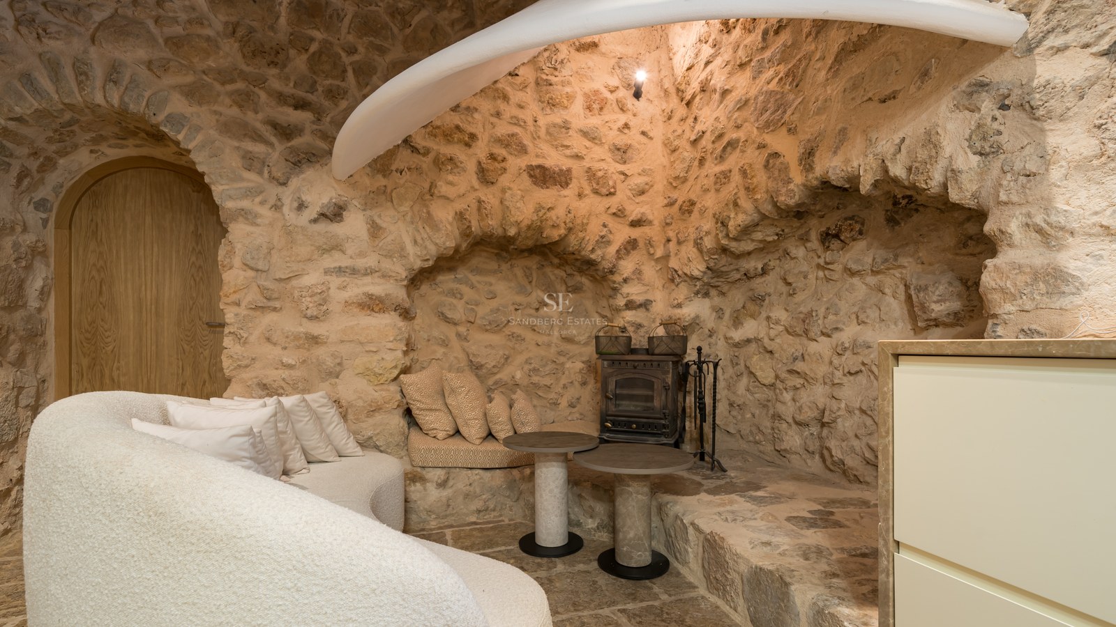 Rustic interior lounge featuring exposed natural stone walls, a stone arch, curved white sofa, and a traditional iron stove.