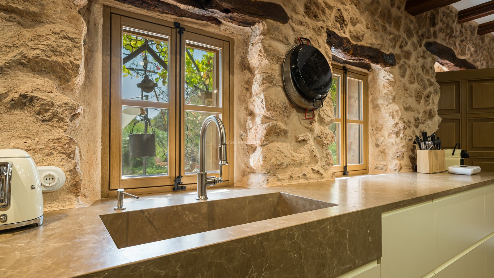 Integrated stone sink under wooden windows in a kitchen featuring exposed stone walls and wooden beams.