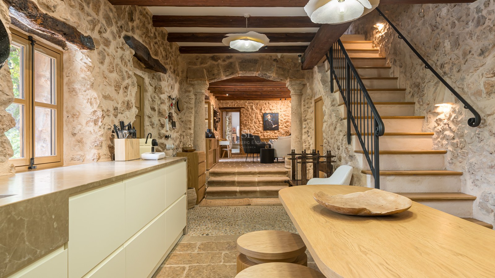 Rustic kitchen with exposed stone walls, wooden beams, a modern island, and a stone staircase leading upstairs.