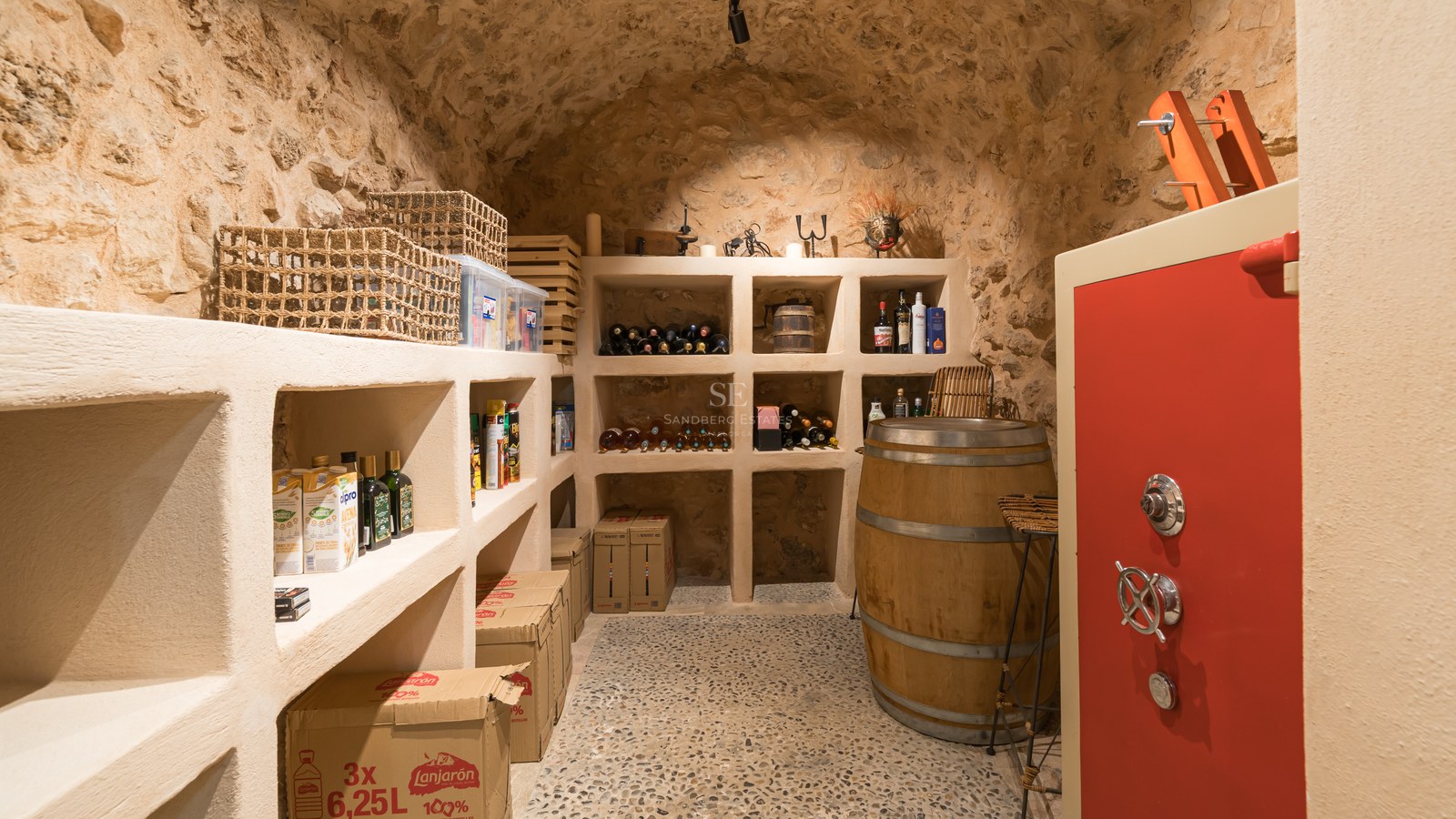 Bodega rústica con techos de piedra abovedados, estanterías de obra, mesa de barril y una caja fuerte roja.