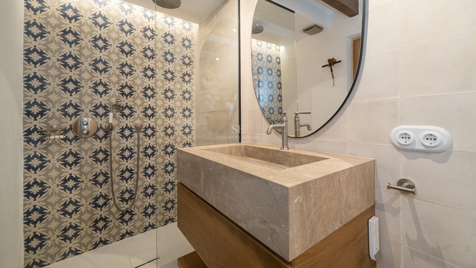 Modern bathroom featuring a large stone vanity, circular mirror, and walk-in shower with blue patterned tiles.