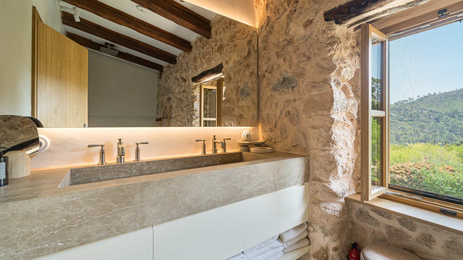 Modern integrated natural stone double vanity against a rustic stone wall with timber beams and a scenic window view.