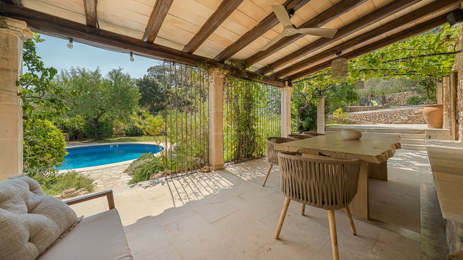 A covered stone terrace with wooden beams, a rustic dining table, and views over a lush garden and turquoise pool.