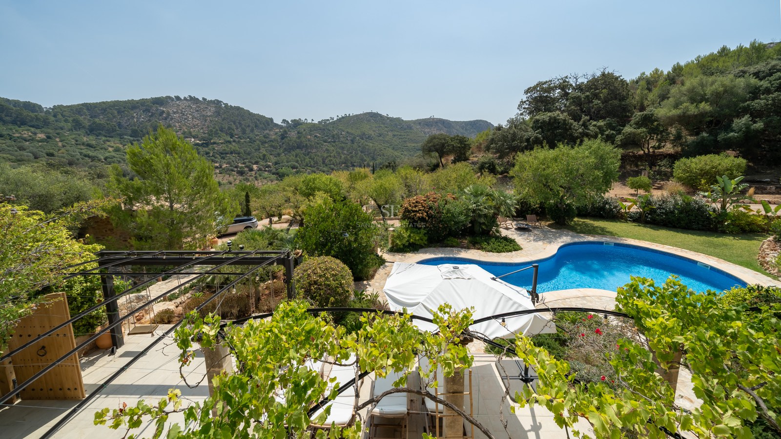 Elevated view of a blue kidney-shaped pool, stone terrace, and lush gardens with green mountains in the distance.