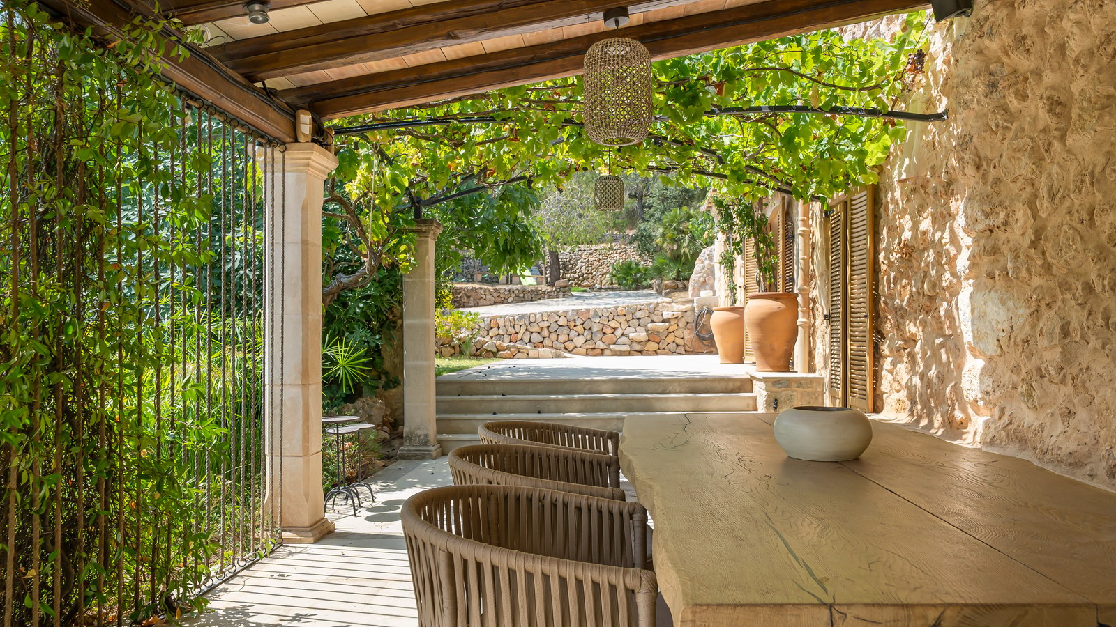 Outdoor dining area under a vine-covered pergola with a live-edge wooden table and natural stone walls.
