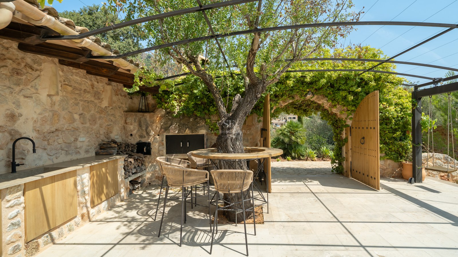 Rustic stone terrace with outdoor kitchen, integrated tree bar table, and arched wooden gates under a metal pergola.